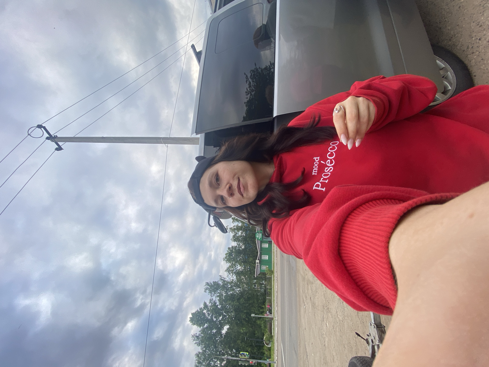 A woman with dark hair, wearing a red hoodie with white text that says "mood Prosecco" and a black baseball cap, takes a selfie. She is standing next to a silver SUV. The background shows a cloudy sky, a utility pole with wires, trees, a traffic light, and a green building. The scene appears to be on the side of a road in the city of Chaikovsky, Russia.