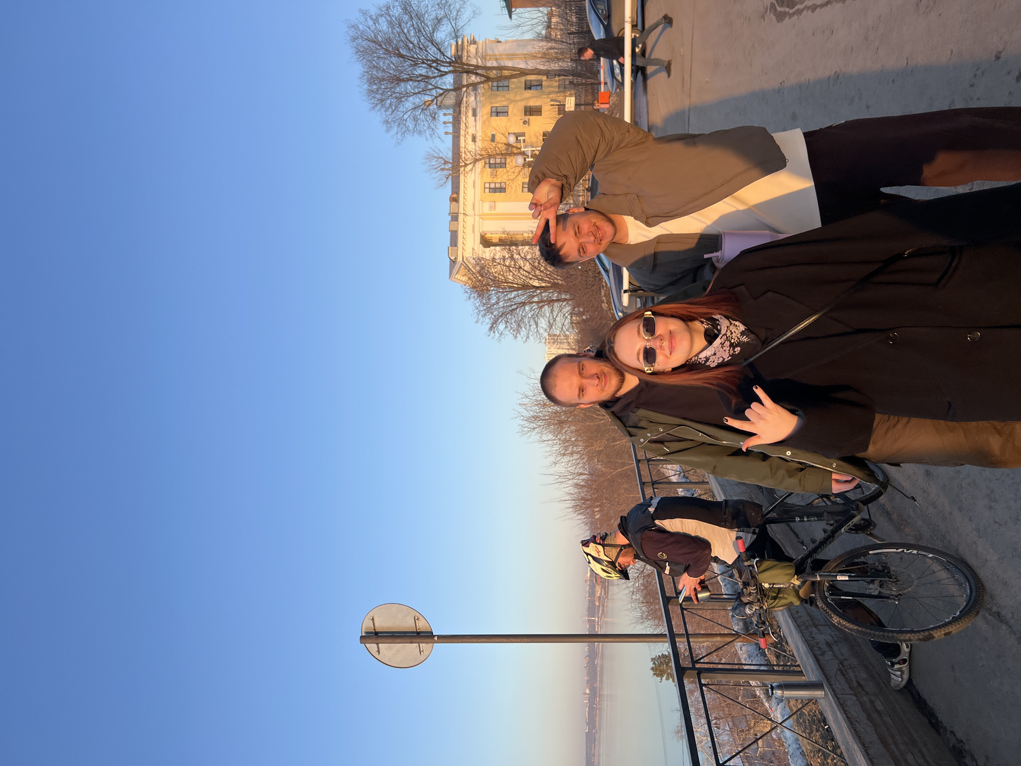 A street scene in Perm, Russia, shows four individuals and a bicycle.
Three people stand close together, facing the camera. A woman with reddish hair wears sunglasses and a black coat. A man with a shaved head stands beside her. A third person, another man, is further back and makes a "peace" sign.
A person in cycling attire stands near a bicycle, looking towards the waterway, while resting a hand on the bridge railing.
The location is a bridge with a metal railing overlooking a river or body of water and land visible in the distance. The sky is blue. A street sign is visible on the left. A building is in the background. Bare trees are also visible.