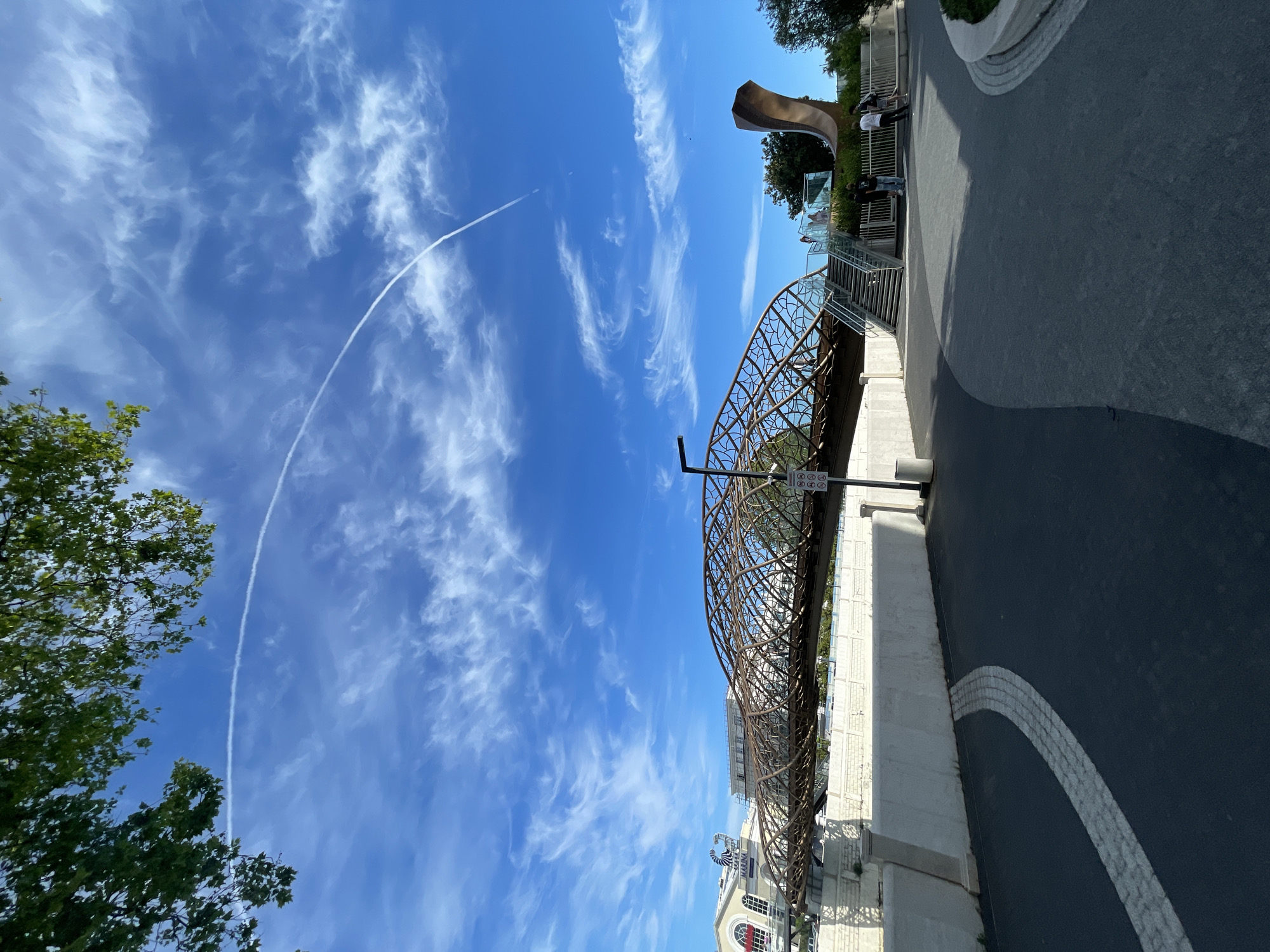 This outdoor photograph captures a vibrant urban scene on a clear, sunny day, likely in Sochi, Russia. The bright blue sky fills the upper half of the image, featuring scattered white cirrus clouds and a prominent, arcing contrail from an aircraft traversing the frame from upper left to lower right.
A distinctive, intricately designed pedestrian bridge, possibly made of bronze-colored metal with a lattice-work pattern, spans the mid-ground. Beneath it, a building with a light-colored facade, arched windows, and the name "MARMALAD" is partially visible.
On an elevated platform to the right, a large, abstract, modern sculpture with a curved, burnished metal appearance stands prominently. Several individuals are present on this platform, some standing near glass railings, others ascending or descending a set of stairs that connect to lower paved pathways. The ground level features dark asphalt paths with decorative white striped curves, bordered by a concrete wall. Green foliage from trees flanks both sides of the scene. A utility pole near the bridge displays a sign with multiple circular symbols, indicative of traffic or public space regulations (though specific text is illegible). The strong, direct sunlight suggests it is midday or early afternoon, making for pleasant conditions in this modern urban landscape.