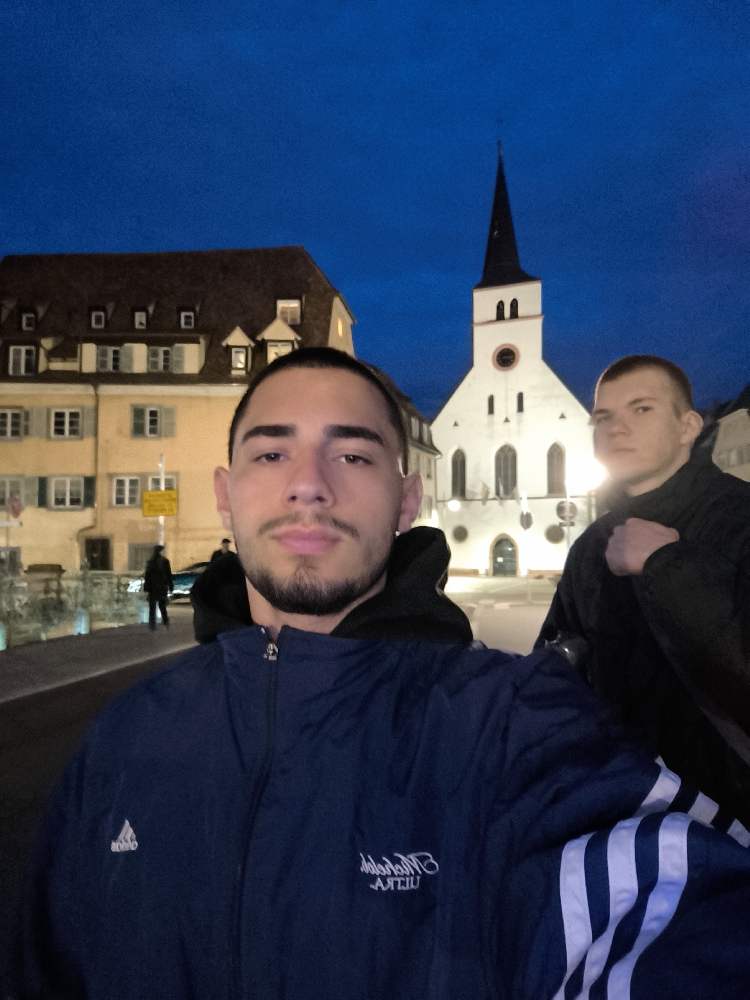The image shows two men standing in front of the Saint-Pierre-le-Jeune Protestant church in Strasbourg, France. The man in the foreground is wearing a blue Adidas jacket. A building with windows and a brown roof is on the left side of the image. The sky is dark blue. There is a street with a person walking in the background.