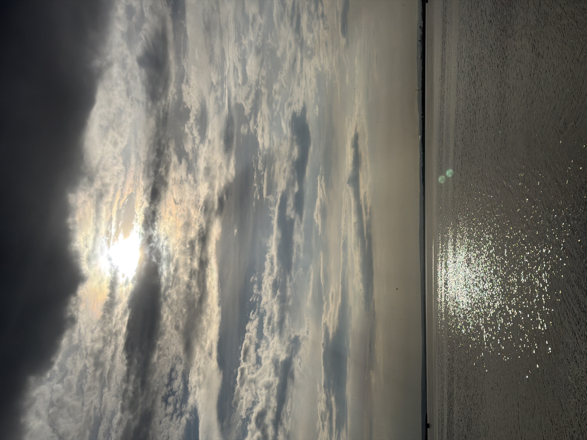 The image shows a body of water, likely the Black Sea, with sunlight reflecting off its surface, creating a sparkling effect. To the left, there is a clouded sky with the sun visible through the clouds. The overall color palette is muted, with shades of gray, white, and touches of yellow and orange from the sun.
The location is Burgas, Bulgaria, as the long strip of coastline is visible across the sea which has buildings on it.