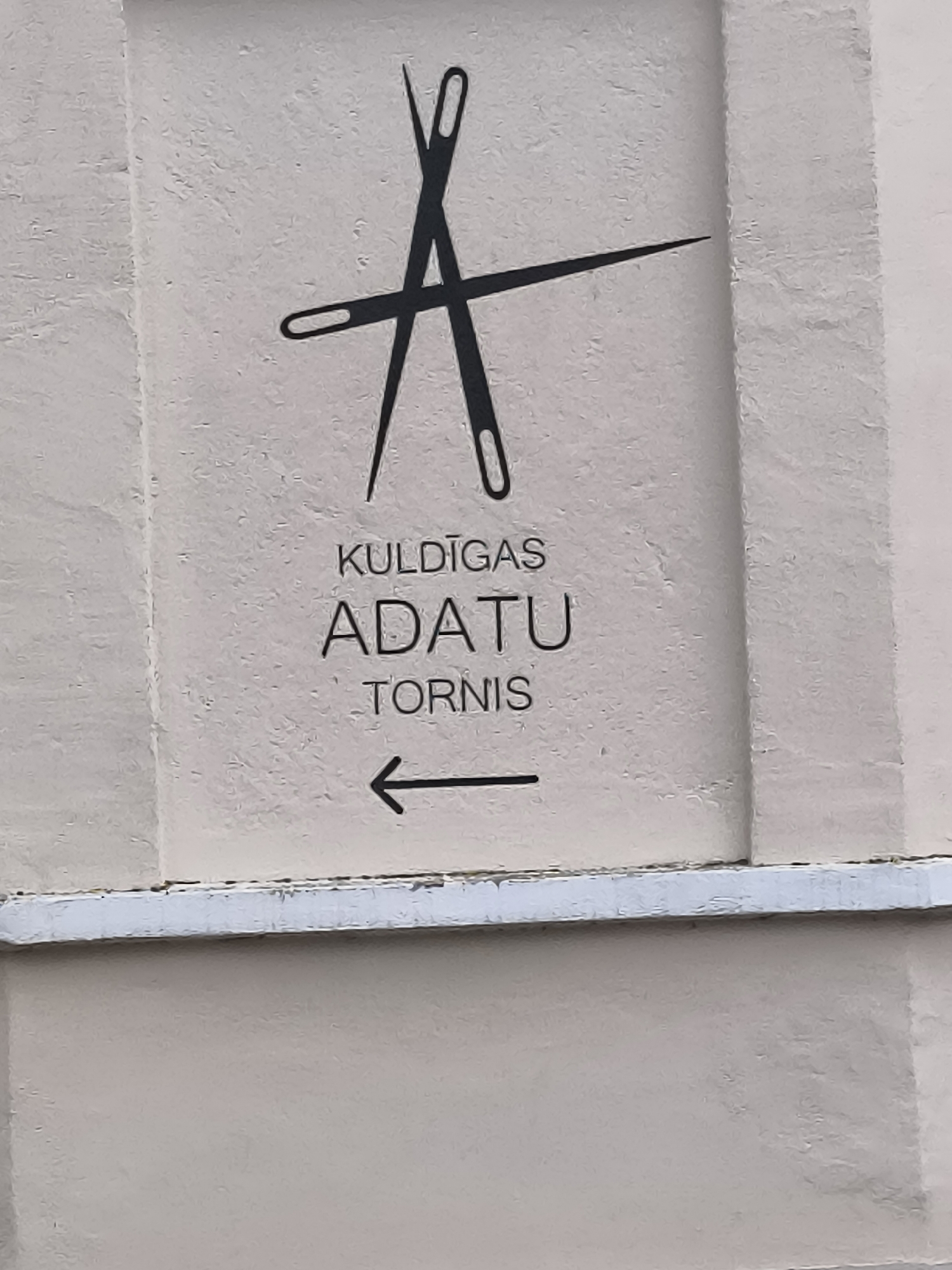 This image shows a sign on a light-colored, textured building wall in Kuldīga, Latvia. The sign features a graphic of two sewing needles crossed in an "X" shape, with one needle pointing upwards and the other downwards. Below the graphic, text reads "KULDĪGAS ADATU TORNIS" (Kuldīga Needle Tower) in a sans-serif font. An arrow pointing to the left is located beneath the text, indicating direction. The building wall appears to be made of stucco or concrete. A white horizontal ledge or trim runs across the lower portion of the image, partially obscuring the lower part of the wall. The lighting suggests an overcast or diffused daylight condition.