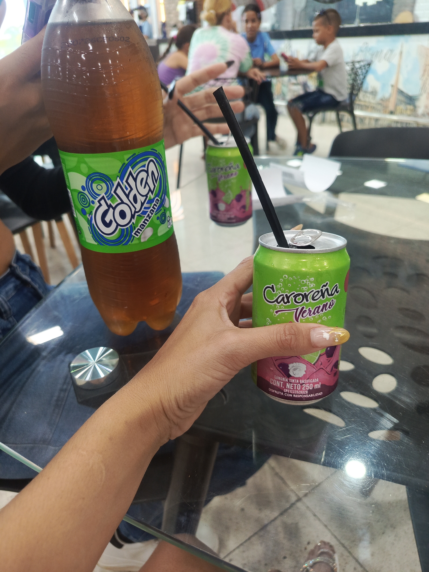 The image shows two drinks on a glass table. A person is holding a green can labeled "Caroreña Verano", and another person is holding an apple flavored soda in a clear bottle with a green label labeled "Golden manzana". Both drinks have straws inserted into them. In the background, people are sitting at tables in an indoor setting. The image was likely taken in Juan Griego, Venezuela.