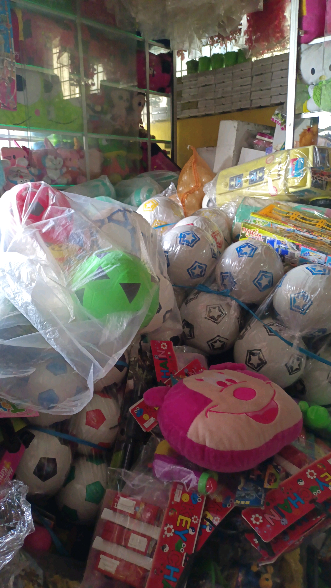 The image depicts a cluttered retail environment, likely a toy store, in Padang Panjang, Indonesia. The foreground is filled with a variety of toys, predominantly soccer balls wrapped in plastic. Several of these soccer balls are white with blue star patterns, while others have less distinct markings. A prominent green soccer ball is visible in the center, also enclosed in plastic. Interspersed with the balls are other toys and merchandise, including a pink plush toy shaped like a character's face, and red packaging with colorful graphics and text, some of which appears to be in Indonesian. In the background, shelves are stocked with more toys, including stuffed animals and various colorful items, some of which are obscured by the foreground objects. The lighting suggests an indoor setting, possibly with natural light filtering in from the rear or sides. The overall impression is one of a busy, perhaps small, retail space specializing in children's toys.
