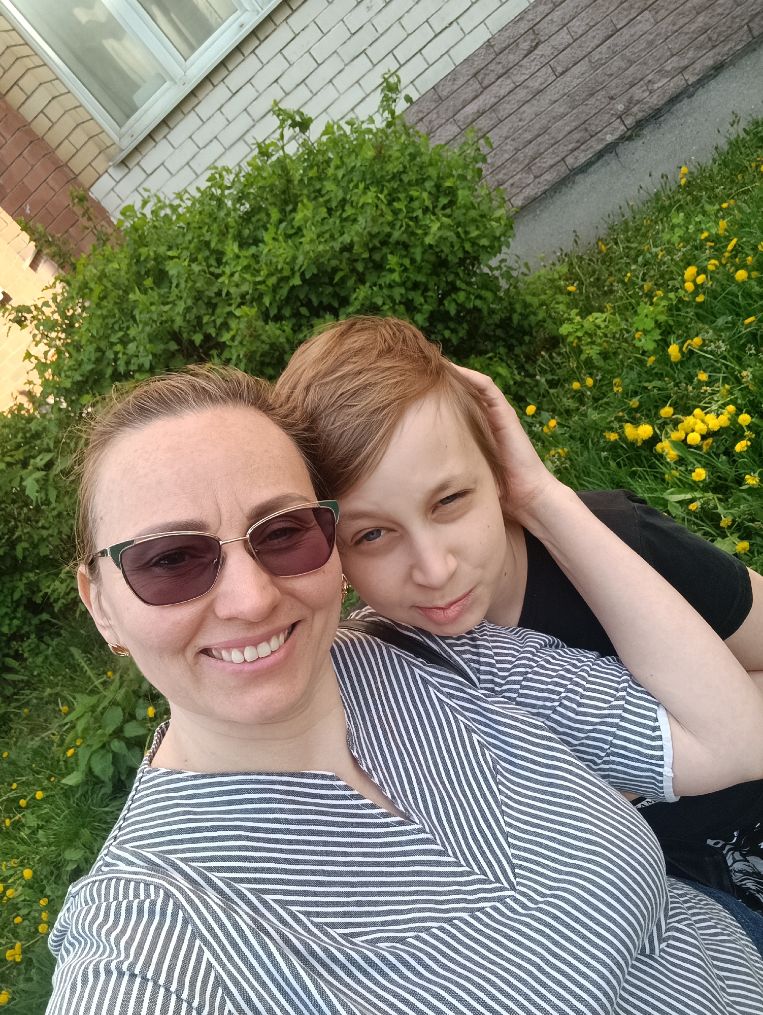 Two individuals, a woman wearing sunglasses and a vertically striped shirt, and a person with short reddish-brown hair in a black shirt, are posing for a selfie outdoors. The woman is smiling and wearing gold earrings. The other person has their hand on their head. The background includes green bushes, grass, and yellow flowers, suggestive of dandelions. A building with brick and white brick exterior is visible behind the vegetation. The location is unknown.