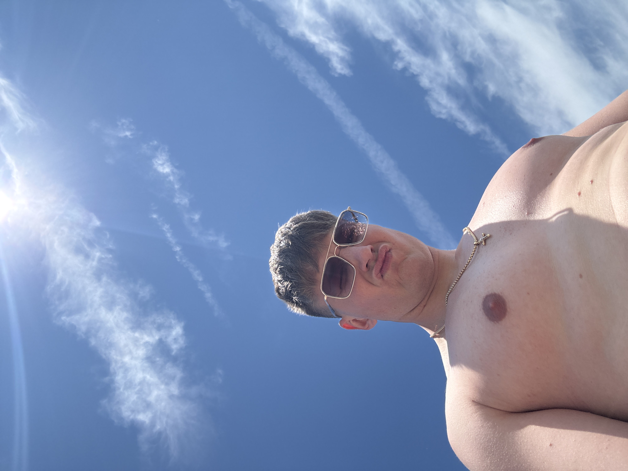 A low-angle shot under bright sunlight shows a shirtless person wearing gold-rimmed sunglasses and a gold cross necklace, with blue sky and wispy clouds as the background. The person's head is tilted, filling the upper half of the frame. The person has short gray hair. The scene is undetermined, but it is within the city of Burgas, Bulgaria.