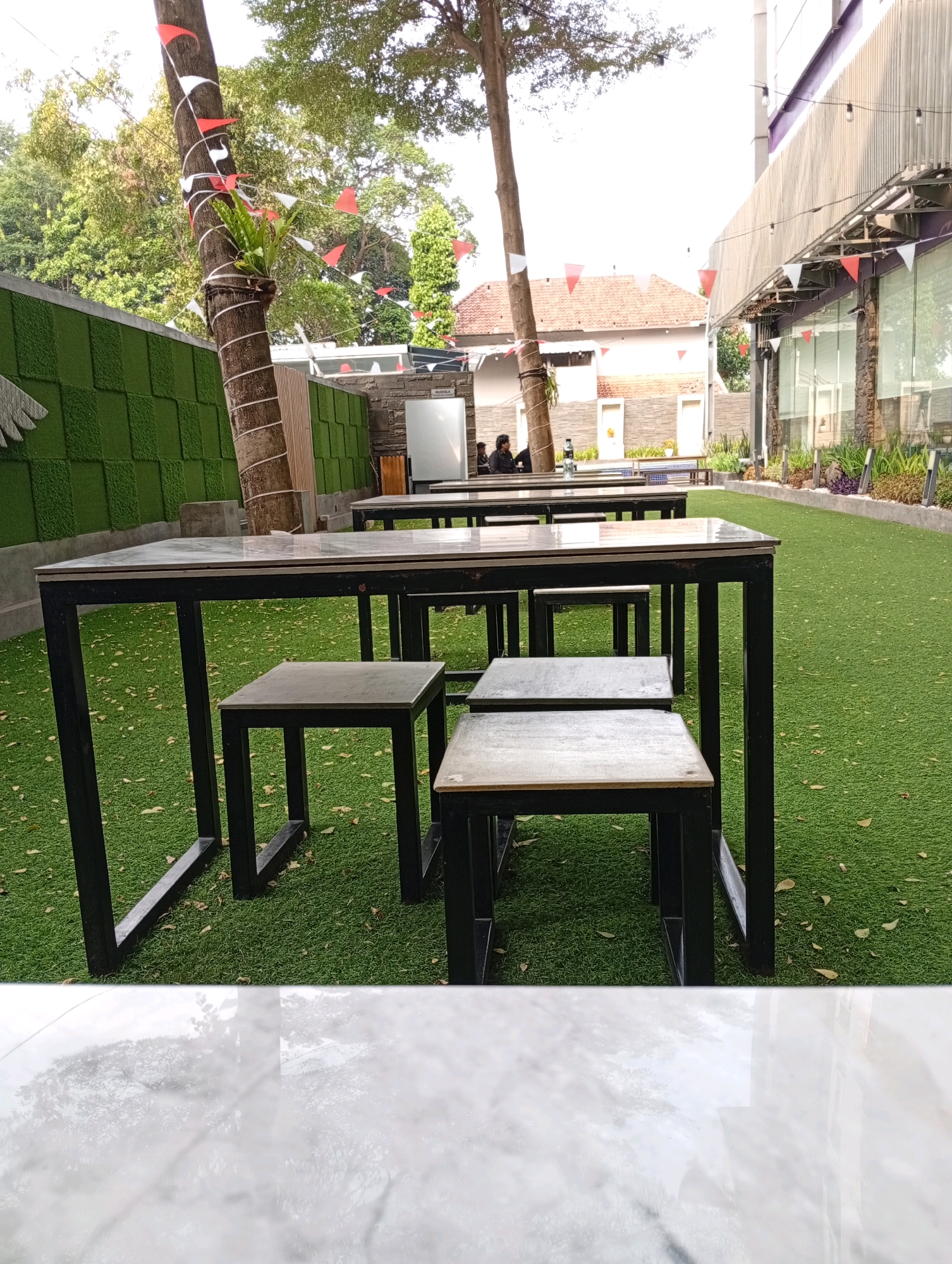 An outdoor seating area in Binong, Indonesia features a row of black metal tables with marble-like tops, accompanied by matching stools. The tables are set on artificial green grass, which is scattered with fallen brown leaves. To the left, a tall tree trunk is wrapped with rope and adorned with small red and white triangular flags, creating a festive atmosphere. Behind the tables, a low green textured wall is visible. In the background, a building with a red tiled roof and a modern building with a corrugated metal facade are present. String lights hang across the facade of the modern building. A few individuals are seated at a table in the distance, engaging in conversation. The foreground is dominated by a reflective white marbled surface.