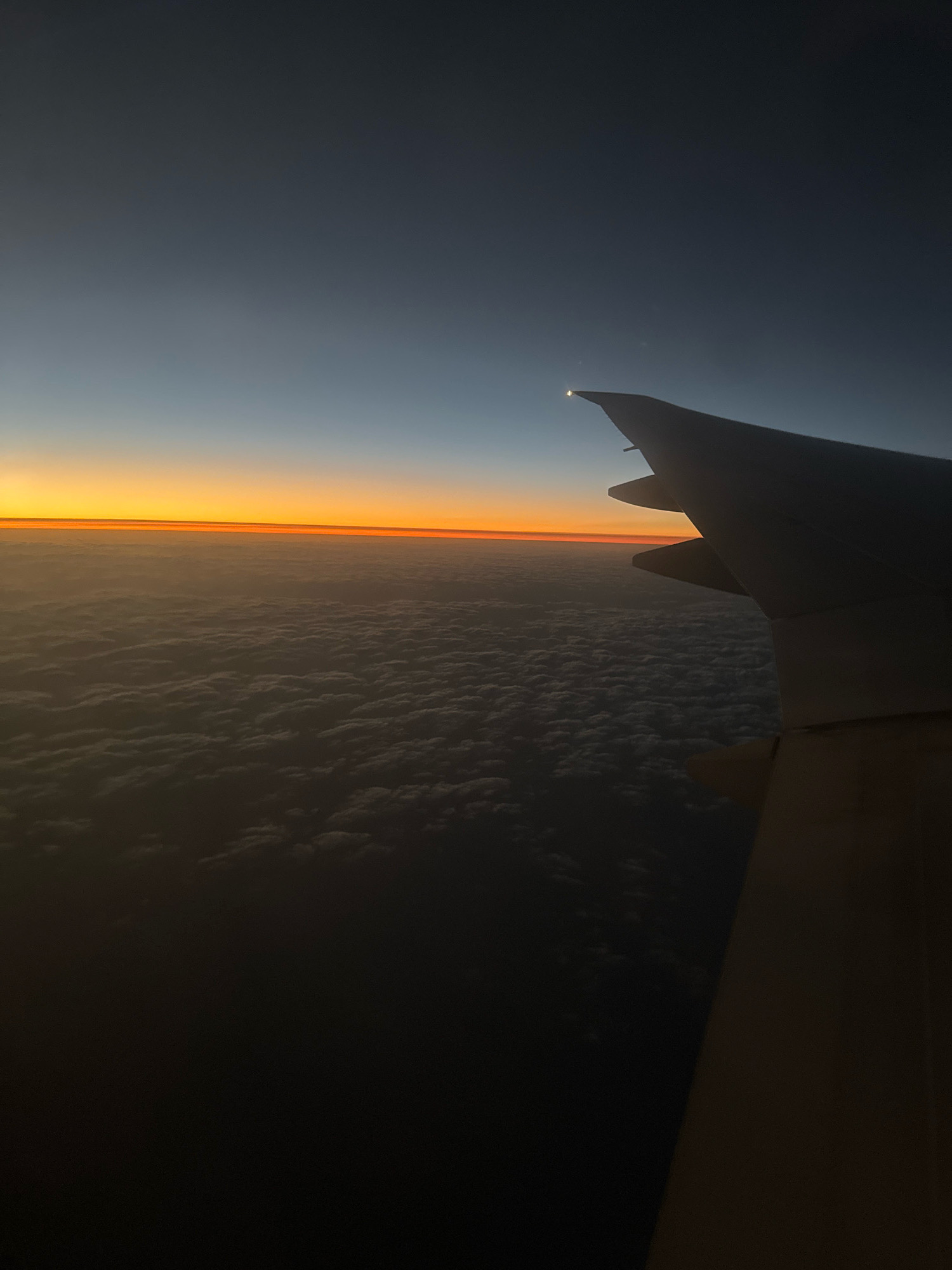 The image is a high-angle shot of an airplane wing against a twilight sky over a cloud-covered landscape. The wingtip, silhouetted against the sky, has a bright light at its apex. The horizon features a thin band of orange and yellow, indicating sunrise or sunset. Below the horizon, clouds are visible, appearing as a textured, white blanket. The sky transitions from a deep gray at the top to a lighter blue near the horizon. The overall scene suggests an aerial view from an airplane in flight. There are no people, specific landmarks of Al Matar, United Arab Emirates, or any discernable actions or interactions. The environment is exclusively the sky, clouds, and an airplane wing.