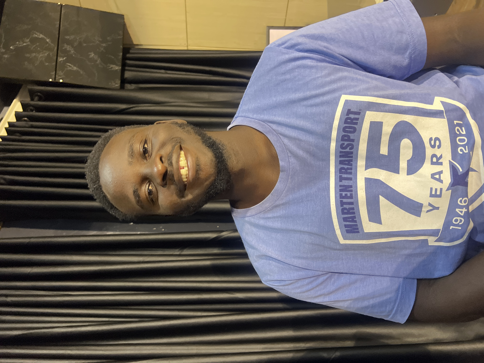 The image shows a medium shot of a Black man wearing a light purple t-shirt with a white and blue graphic. The graphic on the t-shirt reads "MARTEN TRANSPORT 75 YEARS 1946-2021." The man is smiling and looking at the camera. He has short, dark hair and a beard. Behind him is a dark, pleated curtain and a wall with some acoustic paneling. There is no indication of the location being Jos, Nigeria.