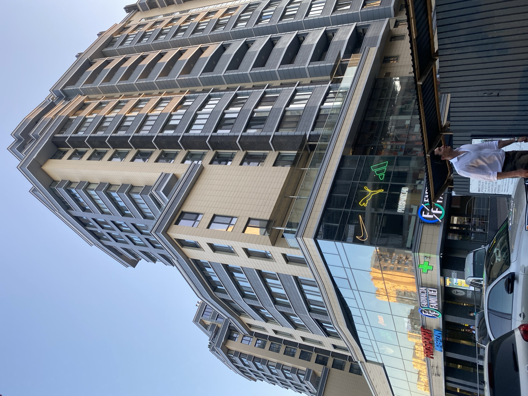 A low-angle shot captures a modern, multi-story building in Chișinău, Moldova, under a clear sky. The building features a beige facade interspersed with gray elements and numerous windows. The lower floors are clad in reflective glass, displaying neon signs for brands such as Nike, Puma, Adidas, and Asics, as well as text identifying "PROSPORT.MD," "FARMARCIA FAMILIEI," and "FARMACIA." A man in a white dress shirt and dark trousers walks towards the right of the frame, partially obscuring a corrugated metal fence. Several cars are parked in front of the building. The scene appears to be a street-level view of a commercial and residential complex.