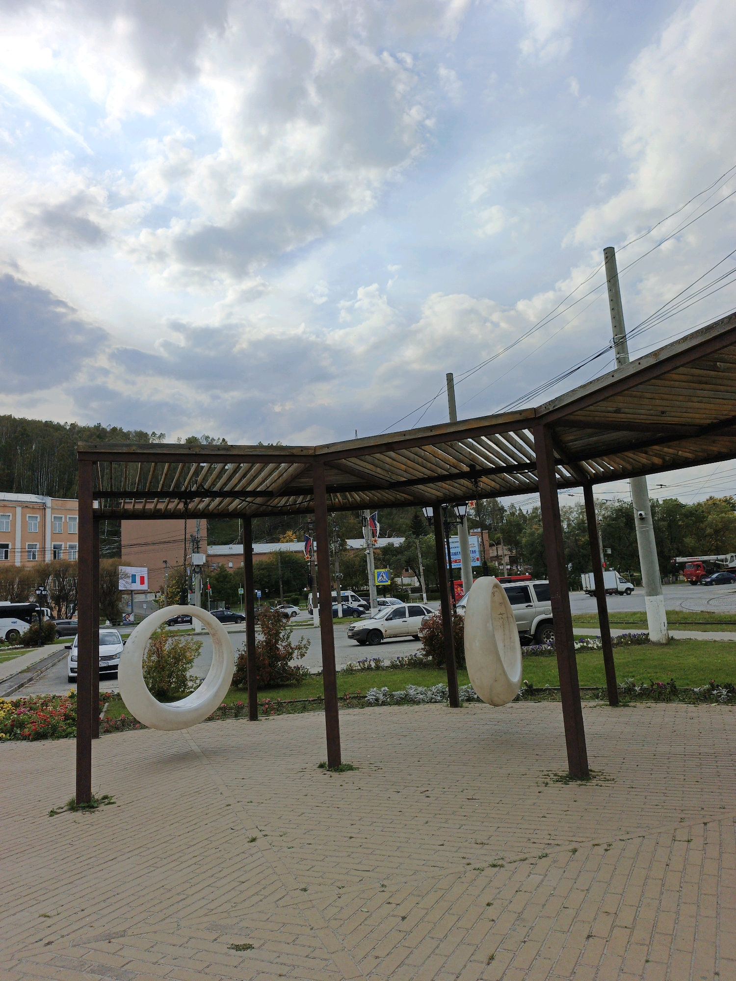 The image depicts a public outdoor seating area with a canopy structure. Two large, circular, white structures, resembling tire swings, are suspended from the canopy by thin wires. The ground is paved with brickwork. In the background, a street with several cars and buildings is visible. The sky is overcast with scattered clouds. The location is identified as the city of Zlatoust, Russia.