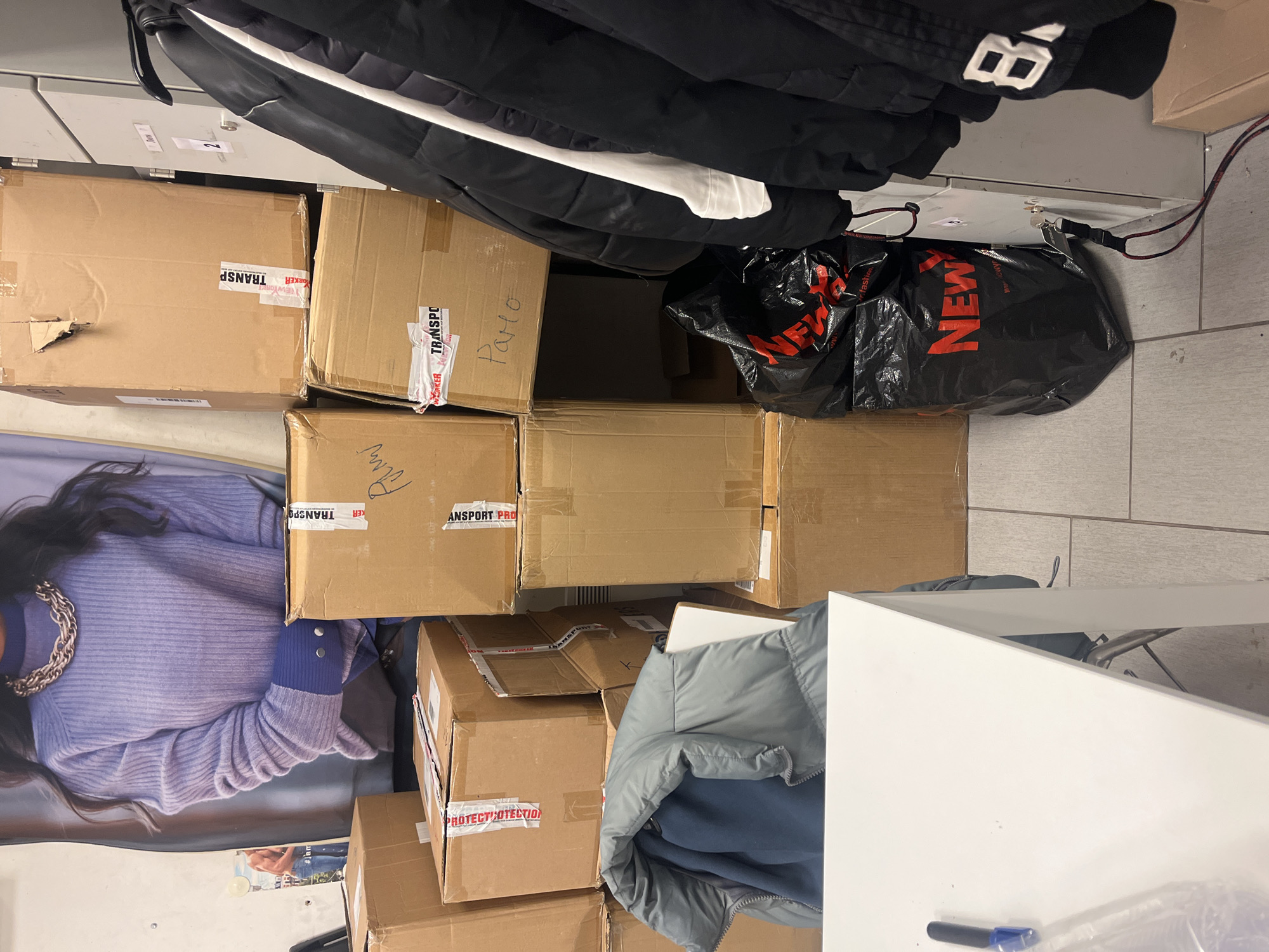 The image captures a cluttered indoor scene. Several cardboard boxes of varying sizes are stacked against a wall, some marked with "TRANSPO" and "PROTECT OTECTIO" tape. One box is labeled "Paro" in handwritten script. A black plastic bag with "NEW" printed in red letters is also present near the top of the stack. Above the boxes hang black jackets, with the number "8" visible on one. To the left, a poster or print of a woman wearing a purple sweater and a large chain necklace is partially visible. In the foreground, a white desk or table is present. There is no explicit evidence to confirm the location is in Sofia, Bulgaria. The scene appears to be a storage area, potentially a stockroom or shipping/receiving department.