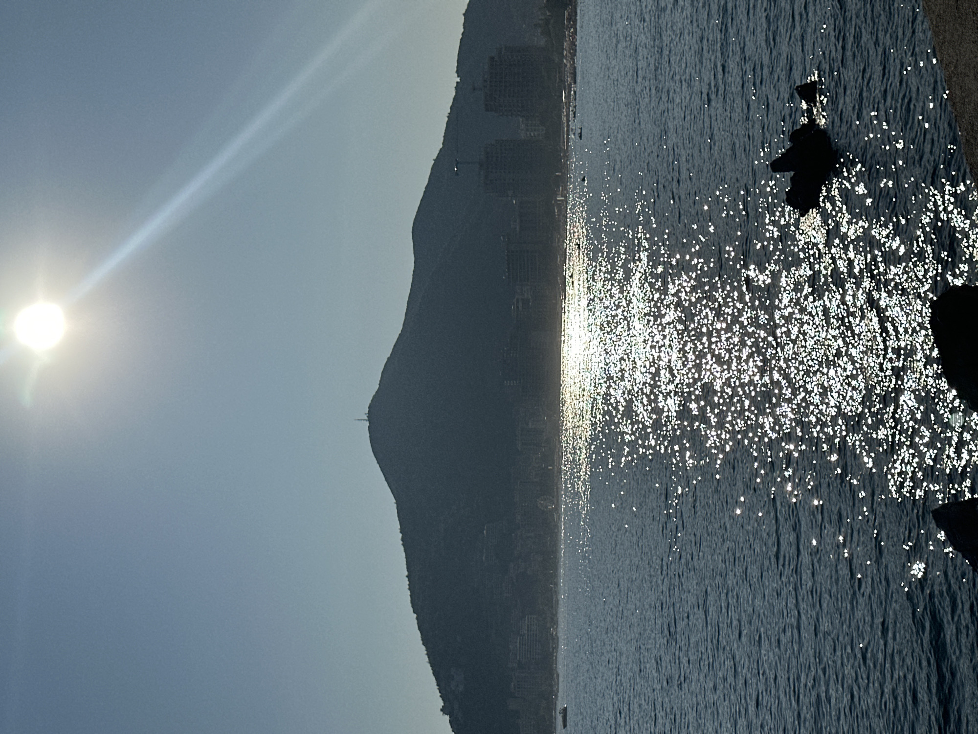 A wide-angle, landscape photograph captures a serene coastal scene in Budva, Montenegro, under a clear, bright sky. The sun, a luminous orb, dominates the upper portion of the frame, casting a radiant, diagonal beam of light across the heavens. Below, a silhouetted mountain range rises majestically, its slopes dotted with faint structures and topped by a prominent antenna. The foothills of the mountain descend towards the coastline, where buildings and the activity of a city are visible in silhouette. The middle ground is occupied by a vast expanse of dark blue sea, its surface shimmering with countless glints of reflected sunlight, creating a dazzling pathway of light from the horizon towards the foreground. The texture of the water is varied, with gentle ripples in some areas and choppier waves in others. In the lower portion of the frame, dark, jagged rocks emerge from the water, adding a dramatic element to the foreground. The overall impression is one of natural beauty and tranquility, with the urban elements of the city subtly integrated into the landscape.