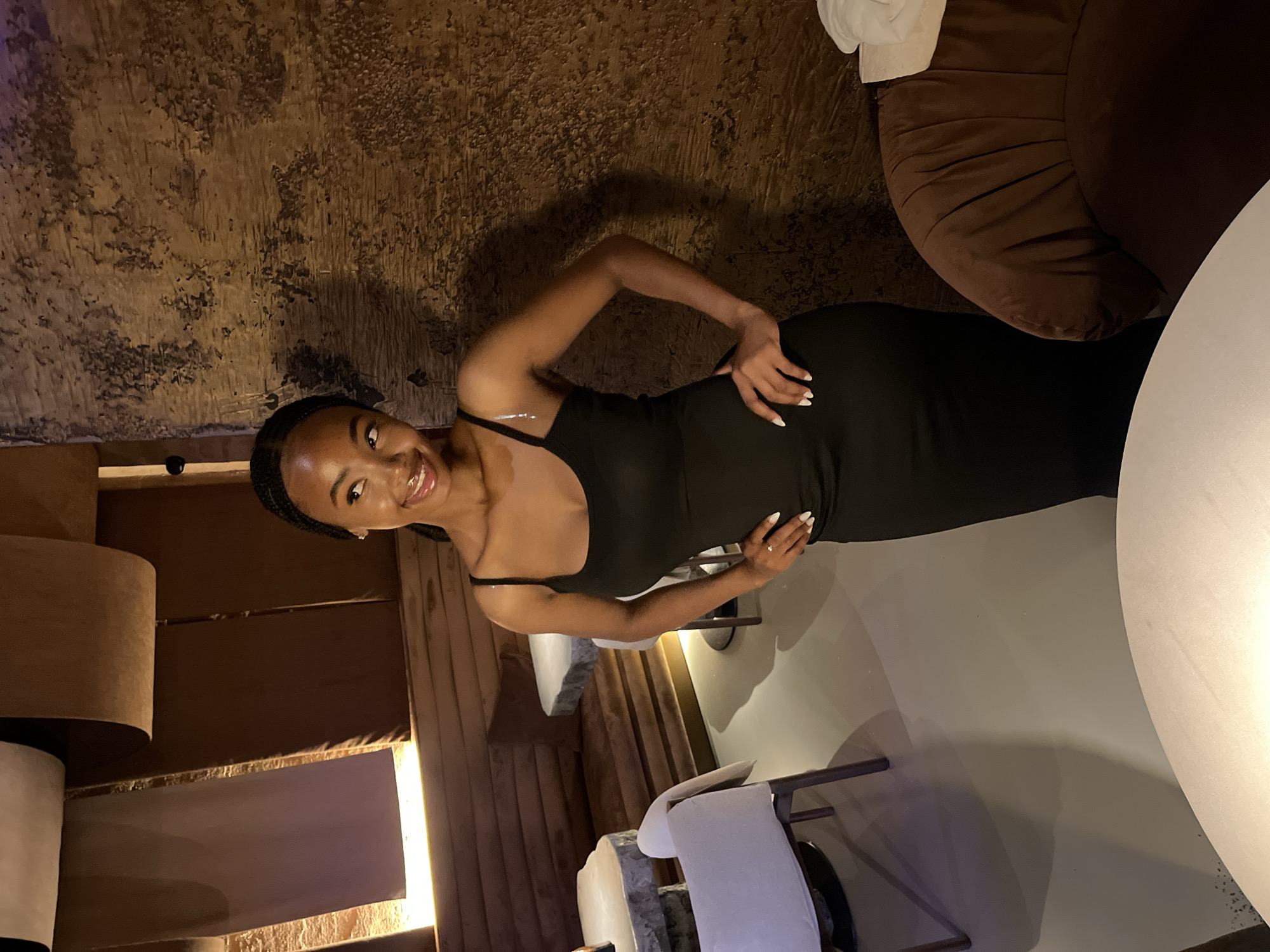 A young Black woman with braided hair and a black sleeveless dress stands with her hands on her hips, looking to her right. She is in a dimly lit restaurant with a textured, dark brown wall behind her. To her left, there are white tables and chairs, and in the background, there are dark brown booths and decorative fabric hanging from the ceiling. The floor is a light gray. The image does not provide any information or context that would allow for determining its location in Kazan, Russia.