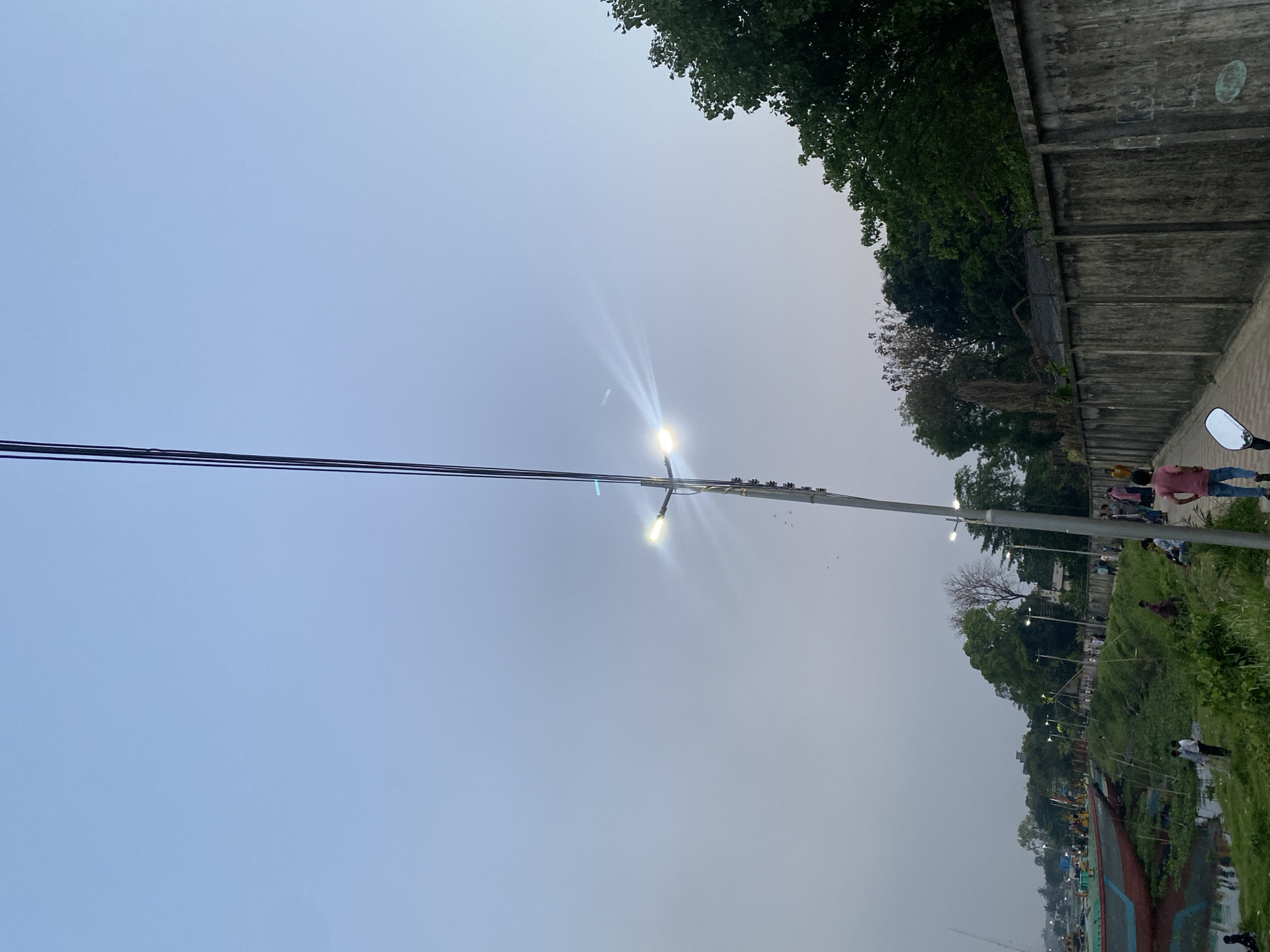 The image depicts a public park scene in Dhaka, Bangladesh. It features a tall, metal light post with multiple lights extending upwards into a cloudy sky. The sky is pale blue, transitioning to a slightly darker shade. Electrical wires run horizontally across the upper portion of the image. A concrete wall and lush trees border the right side of the frame. People are seen walking along a path beside the water. Vegetation grows along the waterfront, and a boat is partially visible in the foreground. The scene suggests a leisurely atmosphere in an urban park setting.