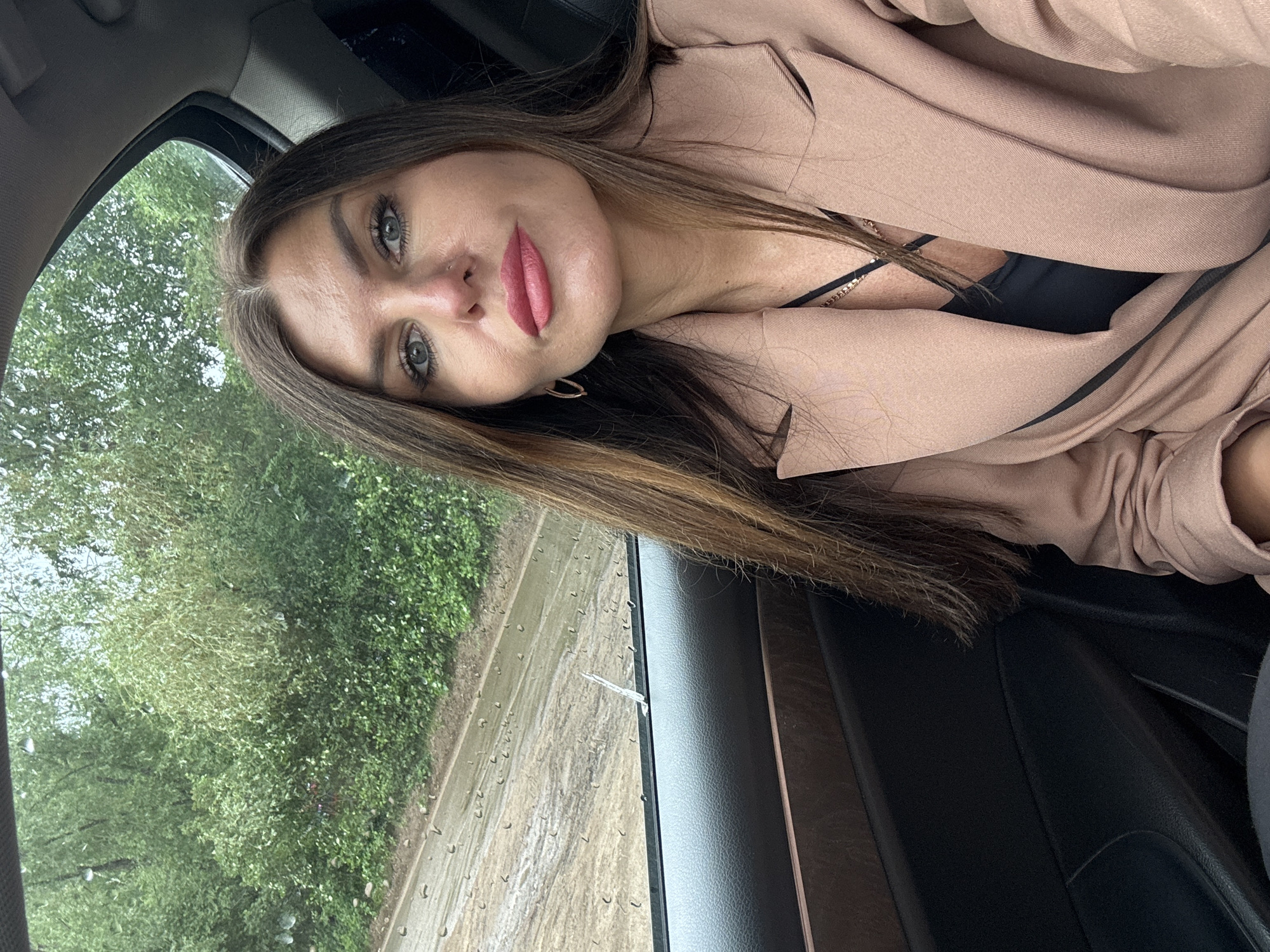 A young woman is captured in a selfie-style photograph, taken from inside a vehicle in Chaikovsky, Russia. She is positioned with her head tilted, looking directly at the camera. She has long brown hair, prominent eyebrows, and striking green eyes. A small stud piercing is visible on her right nostril, and her lips are painted with pink lipstick. She is wearing a light beige or camel-colored blazer over a black top, with a delicate gold-colored chain necklace visible around her neck.
The setting appears to be a car interior, with parts of the dark-colored dashboard and door panel visible to the right and bottom of the frame. Through the car window on the left side, lush green trees and foliage are visible, suggesting an outdoor, possibly natural or suburban, environment. The window itself has numerous raindrops on its surface, indicating that it is raining or has recently rained. Outside, the ground or road appears wet and muddy. The lighting suggests it is daytime, likely overcast due to the rain. There is no visible text in the image.