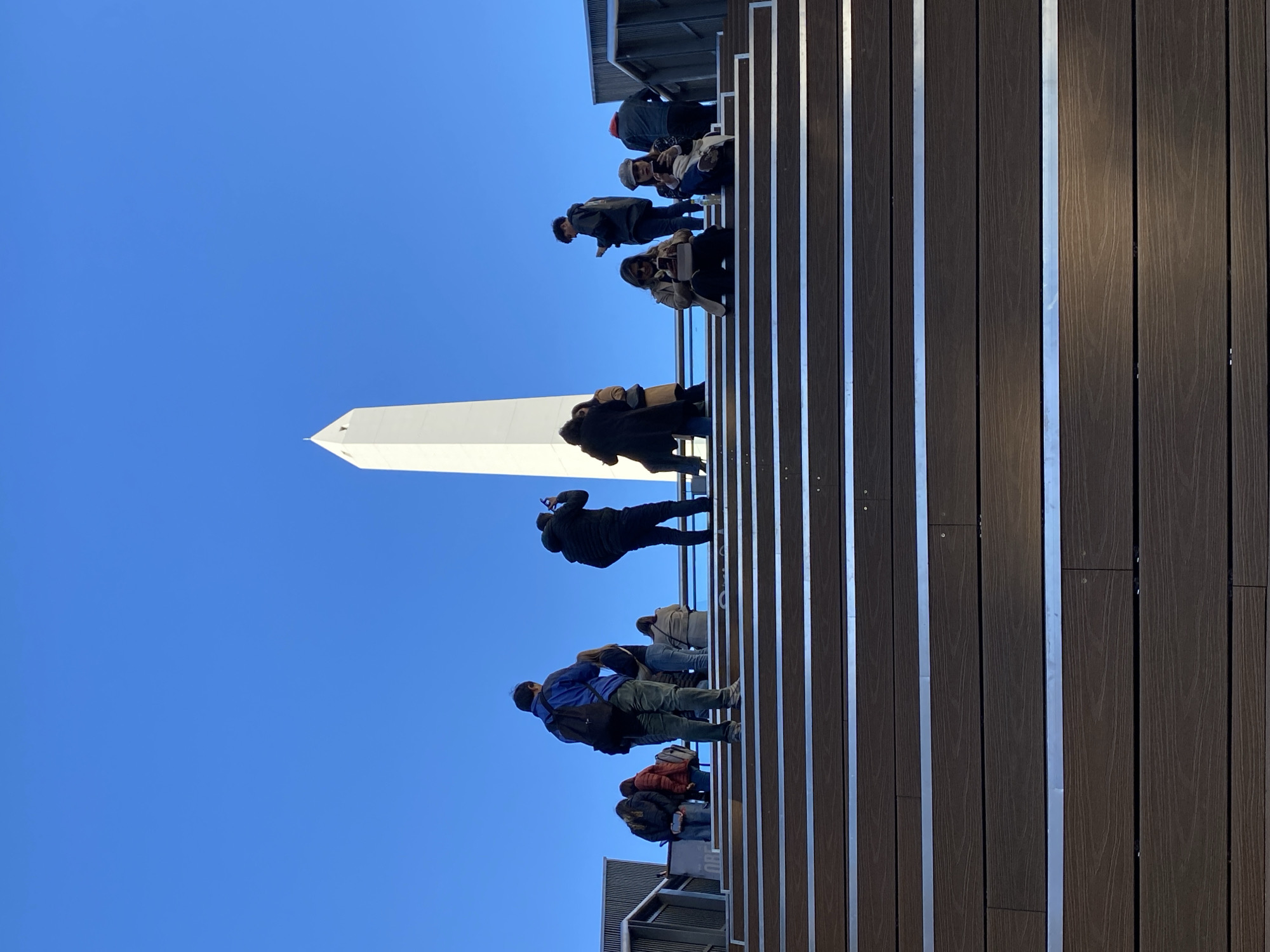 The image shows a group of people standing on a wooden and metal structure against a clear blue sky. In the background is a white obelisk-shaped monument. Based on the monument's appearance and the architectural style of nearby buildings, the location is likely the 1936 Obelisco de Buenos Aires in Plaza de la República, Buenos Aires, Argentina. The individuals appear to be tourists or visitors observing or photographing the monument. They are standing on what appears to be a modern observation platform or walkway.