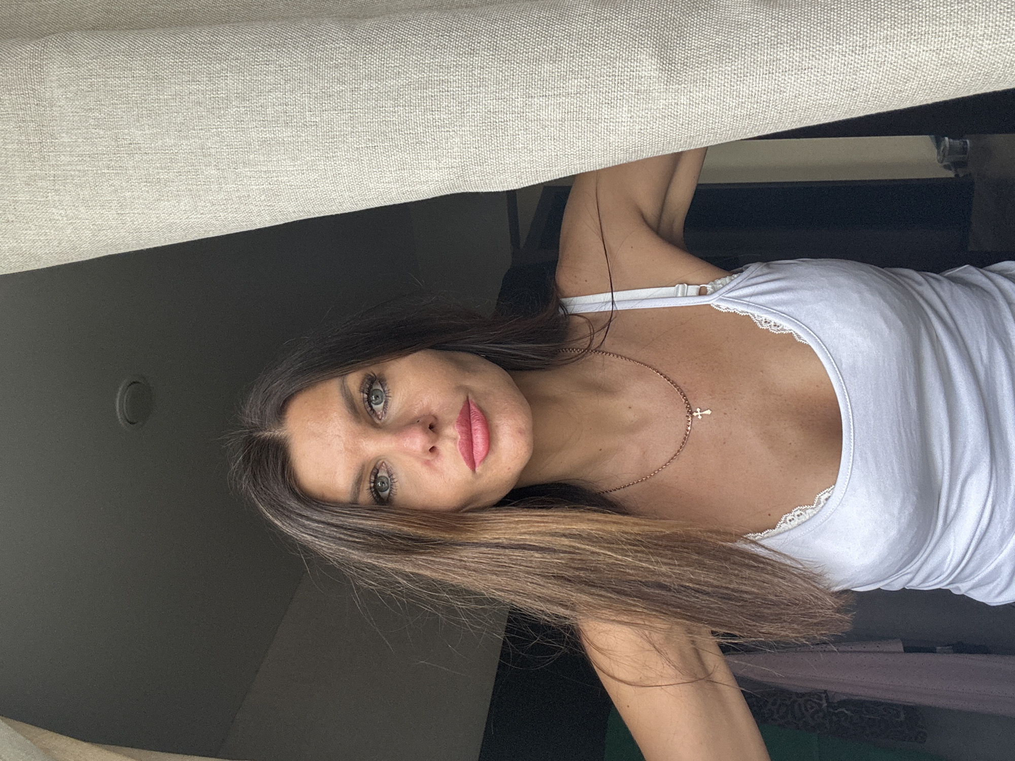 A woman with brown hair and blue-green eyes wearing a white tank top and a gold cross necklace looks at the camera. She is indoors with a beige curtain above her. The background shows glimpses of dark furniture and a wall with a light fixture. The city of Chaikovsky, Russia is not discernible from the image.