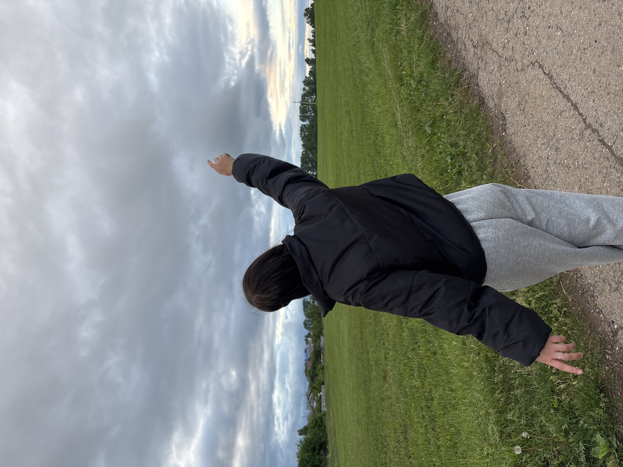 This image depicts a rear view of an individual standing on a paved path adjacent to an expansive green field in Фоки, Russia. The person has dark hair and is dressed in a black puffer jacket and grey sweatpants. Their right arm is raised, with the index finger pointing upwards and slightly to the right, towards the sky. Their left hand is lowered, with the index finger pointing towards the grass; a simple ring is visible on the left ring finger. The foreground features a grey, cracked asphalt path on the right, bordering a vibrant green grass field on the left. Scattered dandelions are visible in the grass in the lower-left corner. In the background on the left, a cluster of trees partially obscures multiple residential structures, indicating a village or settlement. On the right, a distant line of trees stretches across the horizon, with a utility pole visible in the mid-distance. The sky is overcast with multiple layers of grey clouds, displaying a faint yellowish-orange tint on the far right horizon.