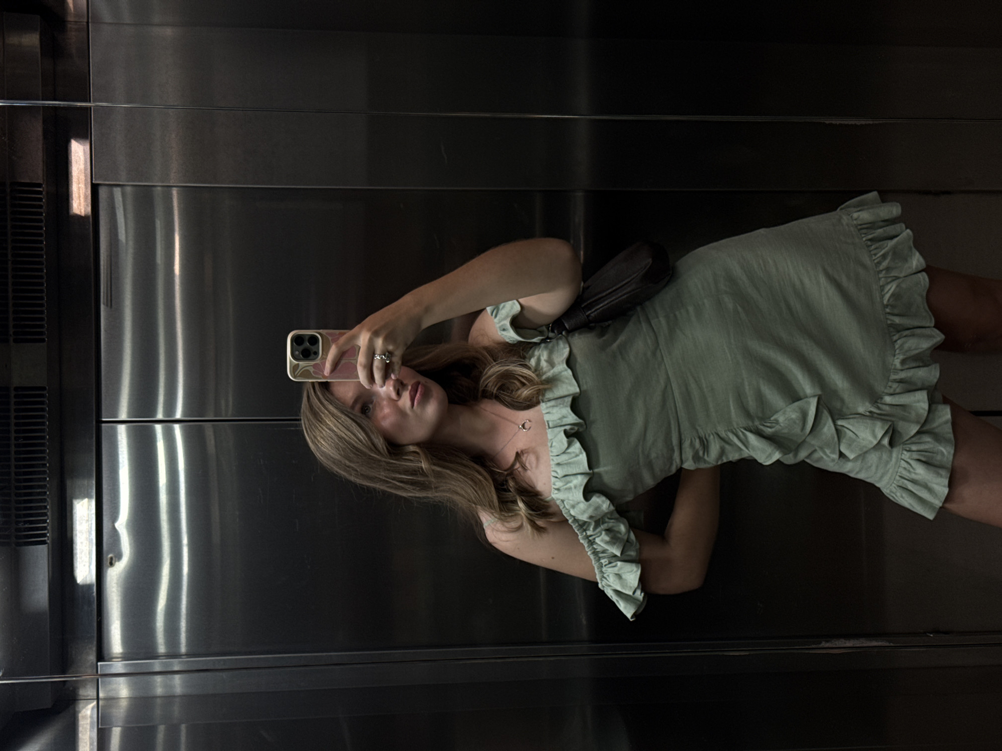 This full-length mirror selfie, taken in the city of Budva, Montenegro, depicts a light-skinned woman inside a brushed metal elevator. The woman, positioned centrally, holds a light-colored smartphone with a visible multi-lens camera module and a patterned case in her left hand, raised to capture her reflection. Her long, light brown hair is parted on the side and falls over her shoulders. She wears an off-the-shoulder, short, ruffled, sage green dress. A silver-toned necklace with a crescent moon pendant is visible around her neck, and a ring is on the index finger of her left hand. A dark-colored shoulder bag is slung over her right shoulder. Her right arm is bent behind her back. The elevator interior features two large, reflective metal panels that form the doors, displaying reflections of overhead lighting and a ventilation grille at the top. The overall lighting is subdued, typical of an indoor elevator environment.