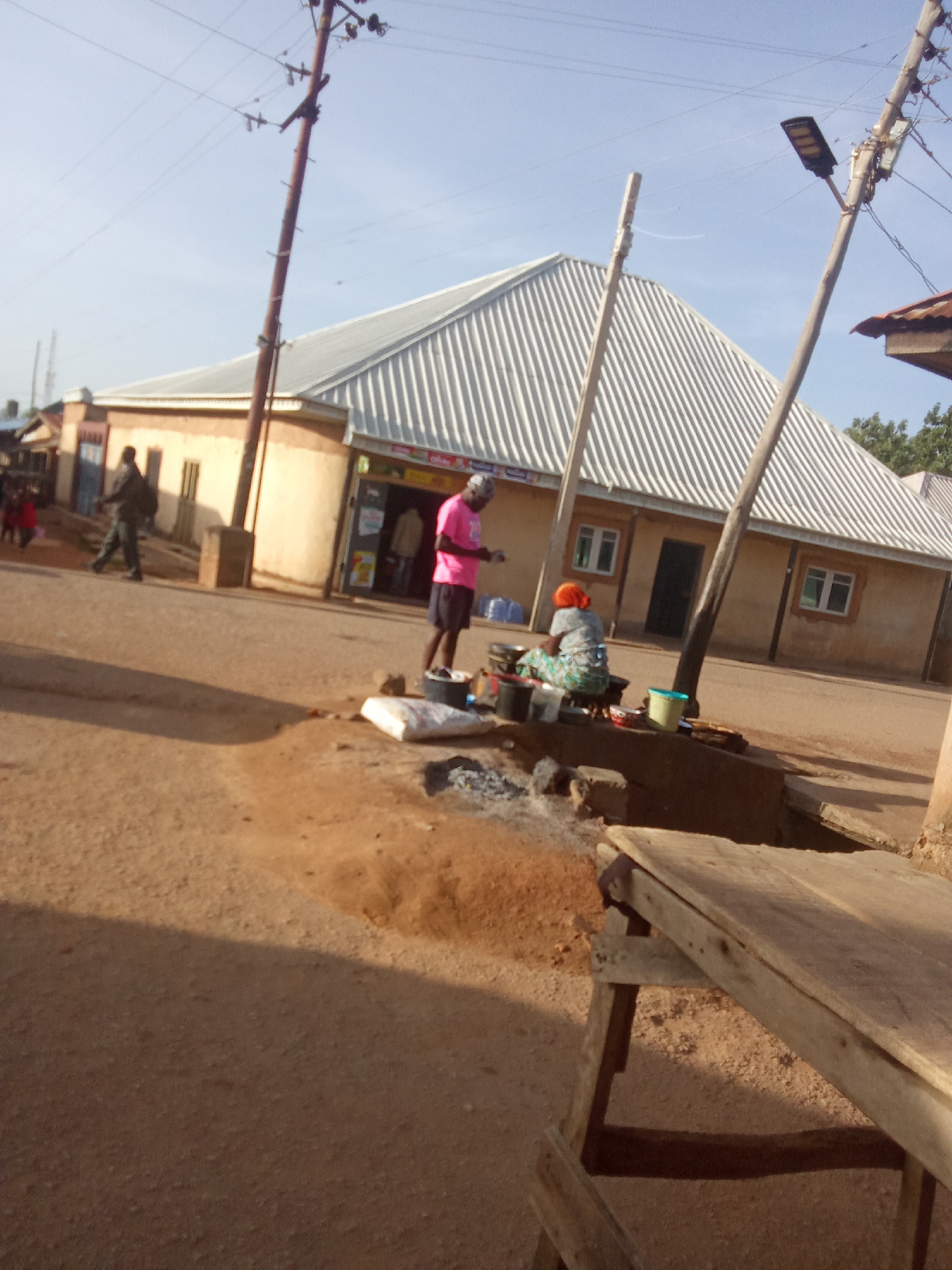 The image depicts a street scene in Jos, Nigeria, under a bright, clear sky. A corrugated metal roof covers a long, single-story building with arched doorways and windows. Several people are visible. In the foreground, a man in a pink t-shirt and dark shorts stands by a small cooking setup on the dirt ground. To his left, a woman in a patterned dress and orange headscarf is seated and appears to be cooking over a fire. A white sack sits near them. In the middle ground, a person in dark clothing walks away from the camera on the dirt road. Further back, a building with an arched entrance is visible, and another individual is seen through the doorway. The street is unpaved, with sand and dirt. A wooden table is positioned in the lower right corner of the image. Utility poles with electrical wires and a street light are present.