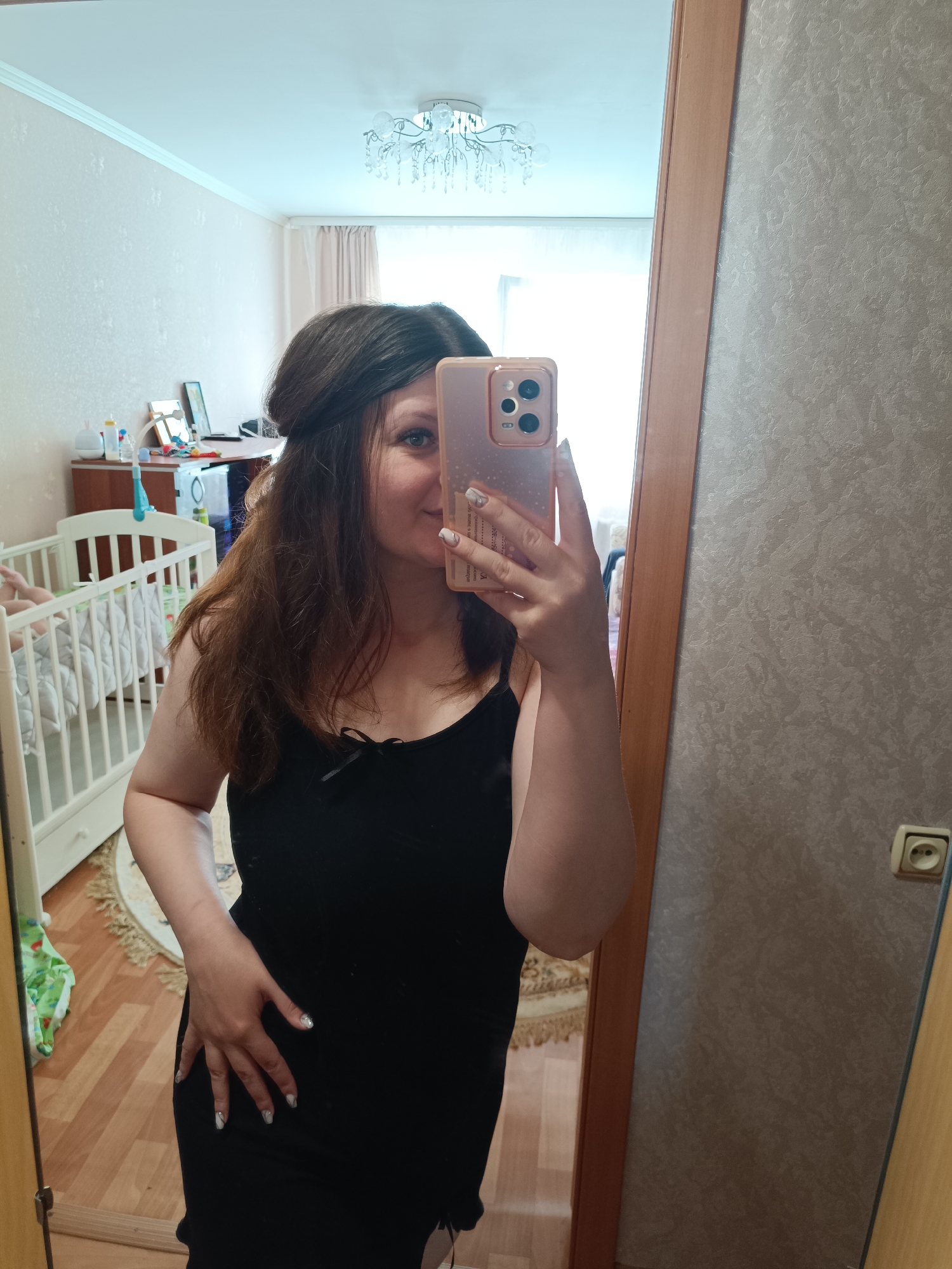 A woman stands in a room, holding a pink smartphone up to her face to take a picture in a mirror. Her dark hair is partially pulled back. She is wearing a black spaghetti-strap top. Her left hand rests on her side. The reflection in the mirror shows a white crib and a dresser in the background. The wallpaper is a light beige with a textured pattern. A light fixture hangs from the ceiling. The wall next to the mirror has a beige outlet. The location is likely Tobolsk, Russia.