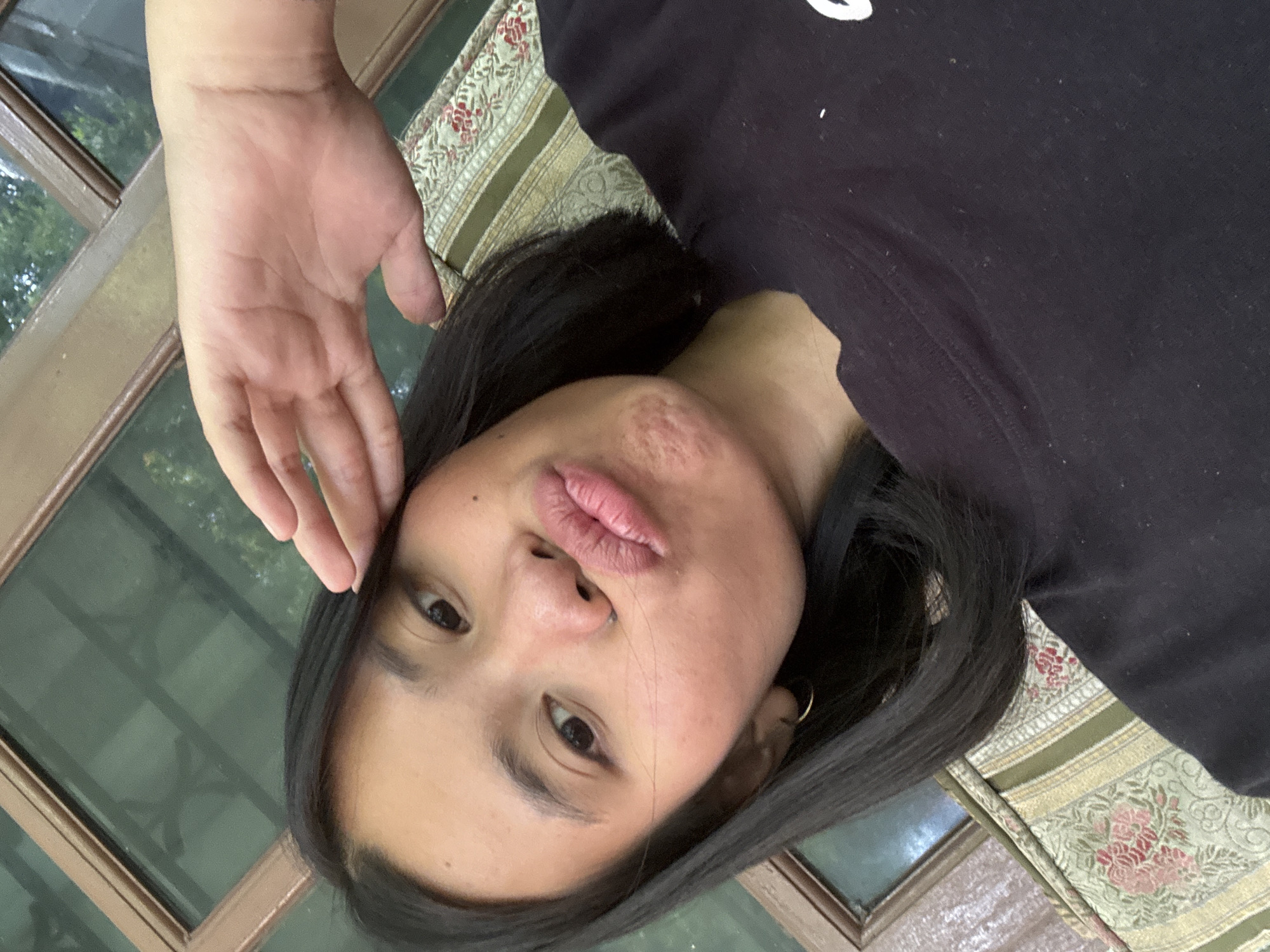 A young woman with dark hair is taking a selfie. She is wearing a black t-shirt and has her right hand raised to her forehead. Her lips are pursed in a pout. She is sitting on a couch with a patterned cushion. The background appears to be a window with wooden frames, possibly in a residential area of South Jakarta, Indonesia. The lighting suggests it is daytime.