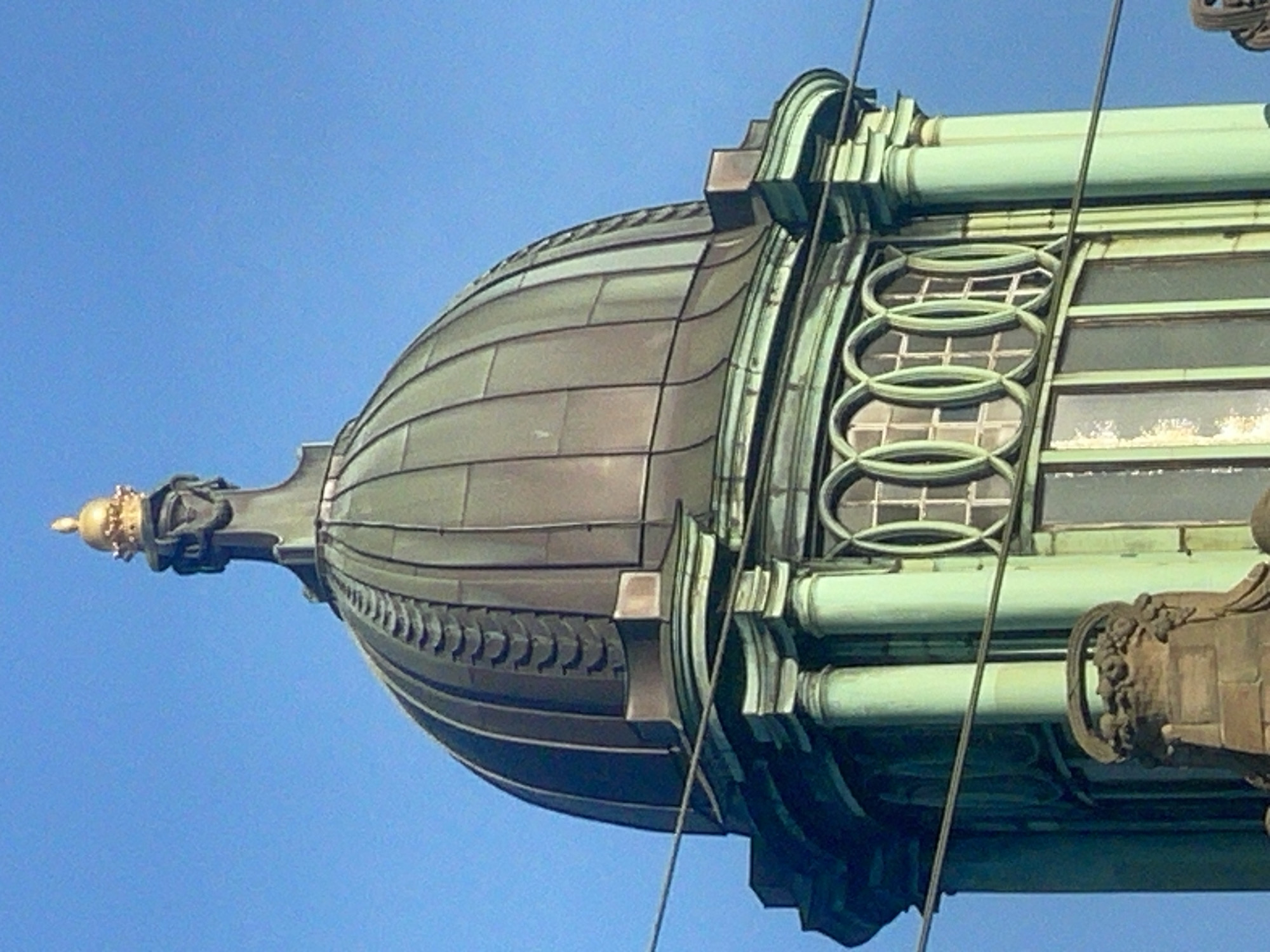 The image shows the top portion of a grand building with a green dome and intricate architectural details, set against a clear blue sky. The dome is segmented and capped with a golden spire. Below the dome, there are several decorative columns painted in a muted green color. A series of oval windows with intricate tracery are visible on the facade, as well as some larger rectangular windows. Decorative stone carvings are present at the base of the visible structure, and two thin wires stretch across the foreground, obscuring parts of the building. The architecture suggests a historical and possibly public building. The location is the Volme Gallery (Volme Galerie) in Hagen, Germany, which is part of the historical building formerly known as the Postamt am Friedrich-Ebert-Platz.
