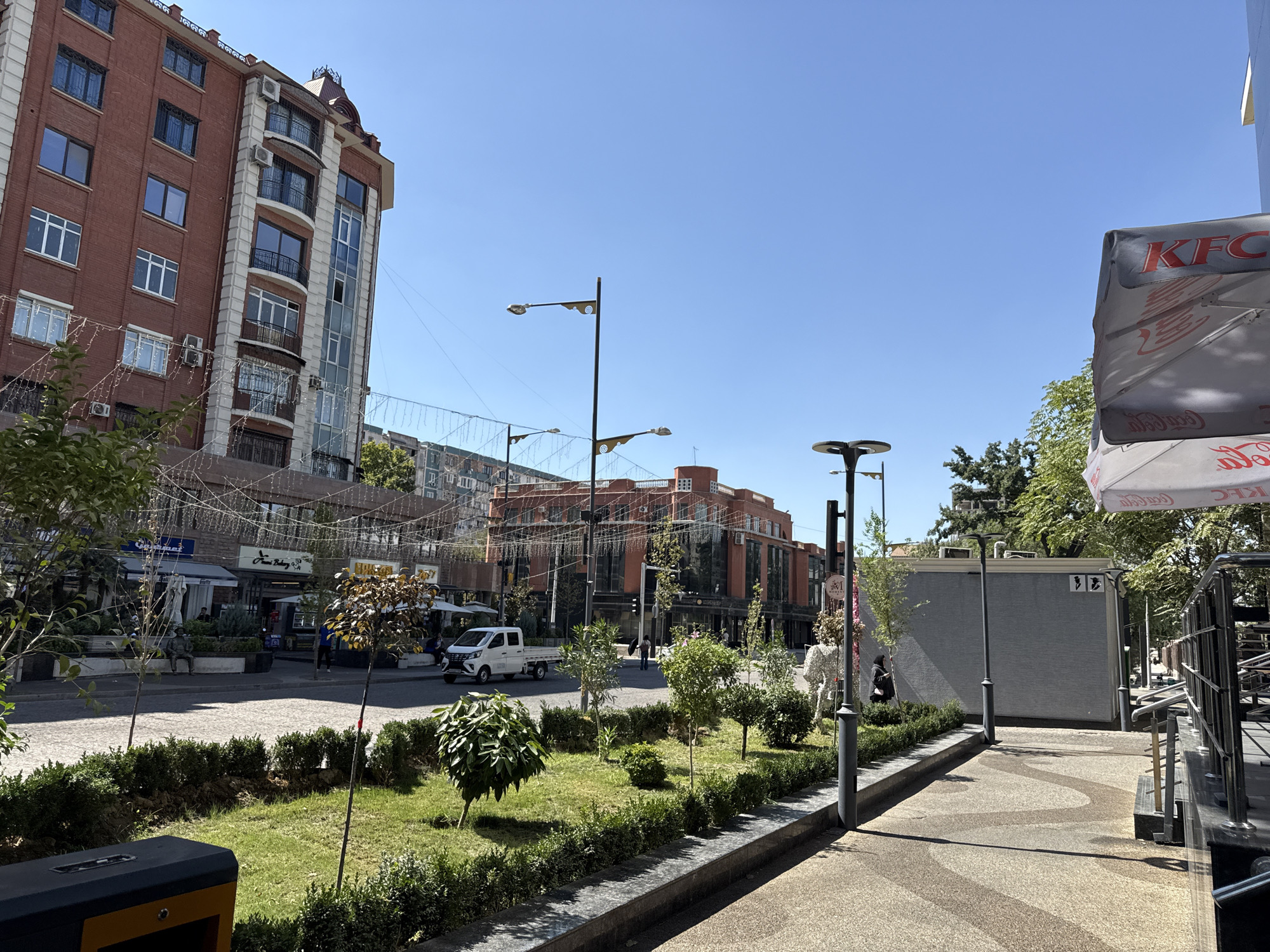 The image depicts a street scene in Tashkent, Uzbekistan, specifically along Amir Temur Avenue, in front of the "Next" shopping mall. In the foreground, a green strip with manicured bushes and trees separates the sidewalk from the street. A dark rectangular bin is visible on the left. The street is paved with cobblestones, and a white truck is parked on the right side of the road.
Across the street, several multi-story buildings line the avenue. The building on the left is a red brick structure with white decorative elements and numerous windows. A taller, modern building with a glass facade is adjacent to it. Further down the street, another brick building with a modern architectural style is visible. Shops and cafes are identifiable at street level, with awnings and umbrellas providing shade. Decorative lights are strung between buildings across the street.
Several people are present. Two individuals are walking on the sidewalk across the street. A person is seated on a bench in a landscaped area in front of a shop. Another individual is walking towards the right side of the frame near a grey building. On the right side of the image, part of a KFC umbrella is visible, suggesting the presence of a fast-food establishment. The sky is clear and blue, indicating a sunny day.