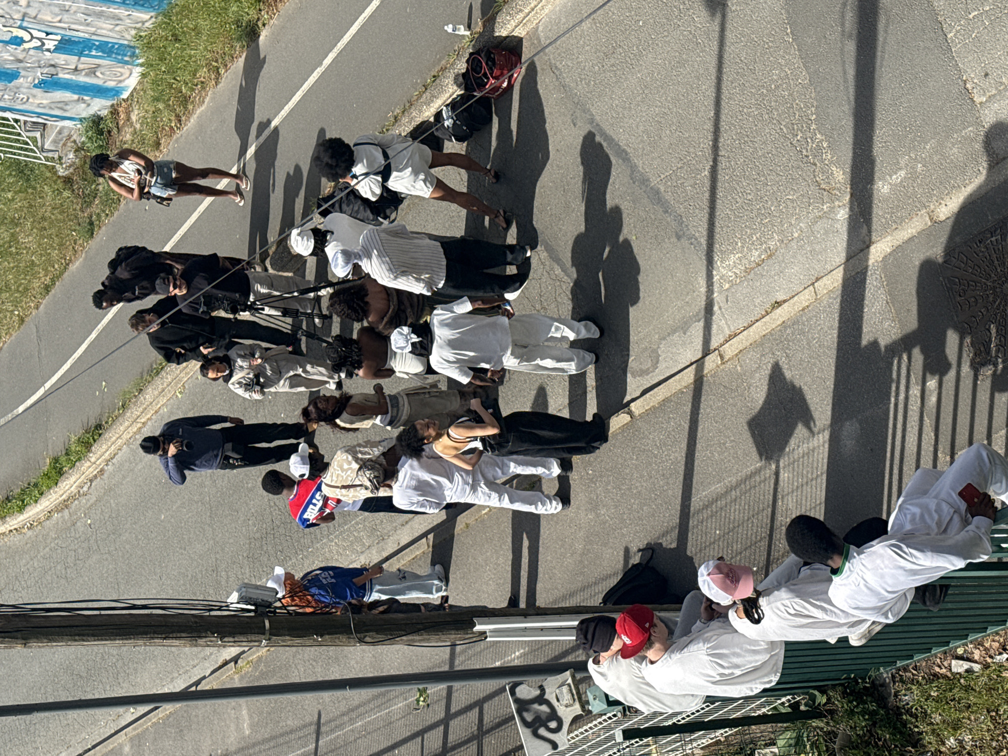 An overhead, high-angle view captures a group of approximately fifteen to twenty people gathered on a sunny street and sidewalk in Charenton-le-Pont, France. The scene suggests an outdoor production or filming activity, with several individuals holding professional cameras on tripods.
Many subjects are young adults dressed in casual clothing. Some wear all-white outfits, while others sport various streetwear styles. Notable clothing includes a blue basketball jersey with "BULLS" and the number "23" visible, a camouflage-patterned jacket, and several baseball caps in red, pink, and white, as well as a black beanie. One woman wears a white tank top and denim shorts, holding a camera.
The group is dispersed across an asphalt road and a concrete sidewalk, bordered by a grassy bank and a metal fence on one side, and a concrete wall with blue and white graffiti on the other. Long, distinct shadows indicate bright sunlight, suggesting a clear day during the late morning or early afternoon.
In the foreground, two individuals are seated on a green metal bench, both dressed in white. One holds a red smartphone. Behind them, on a concrete surface, a stylized black snake graphic is visible. Utility poles with wires run across the upper part of the frame. Scattered on the ground near some individuals are bags, including a red backpack and a black duffel bag. A patterned manhole cover is visible on the sidewalk.