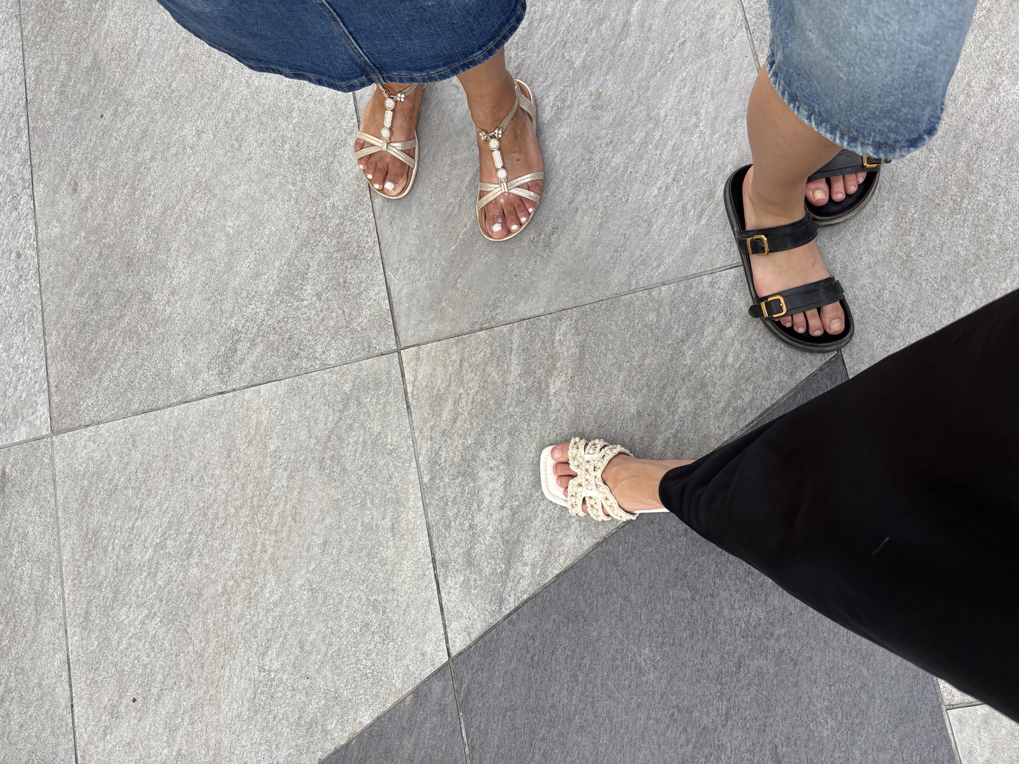 The image captures a ground-level perspective of at least two individuals' feet standing on a tiled surface. One person is wearing black pants and white sandals adorned with pearls and decorative elements. Another person is wearing blue jeans and gold-colored sandals with pearl and bead accents. A third person's feet are visible wearing black sandals with gold buckles and blue jeans. The tiled ground appears to be composed of large, rectangular tiles in varying shades of gray, suggesting an outdoor urban environment. The scene is devoid of any specific landmarks that would pinpoint a location in Orihuela, Spain. The lighting suggests it is daytime.