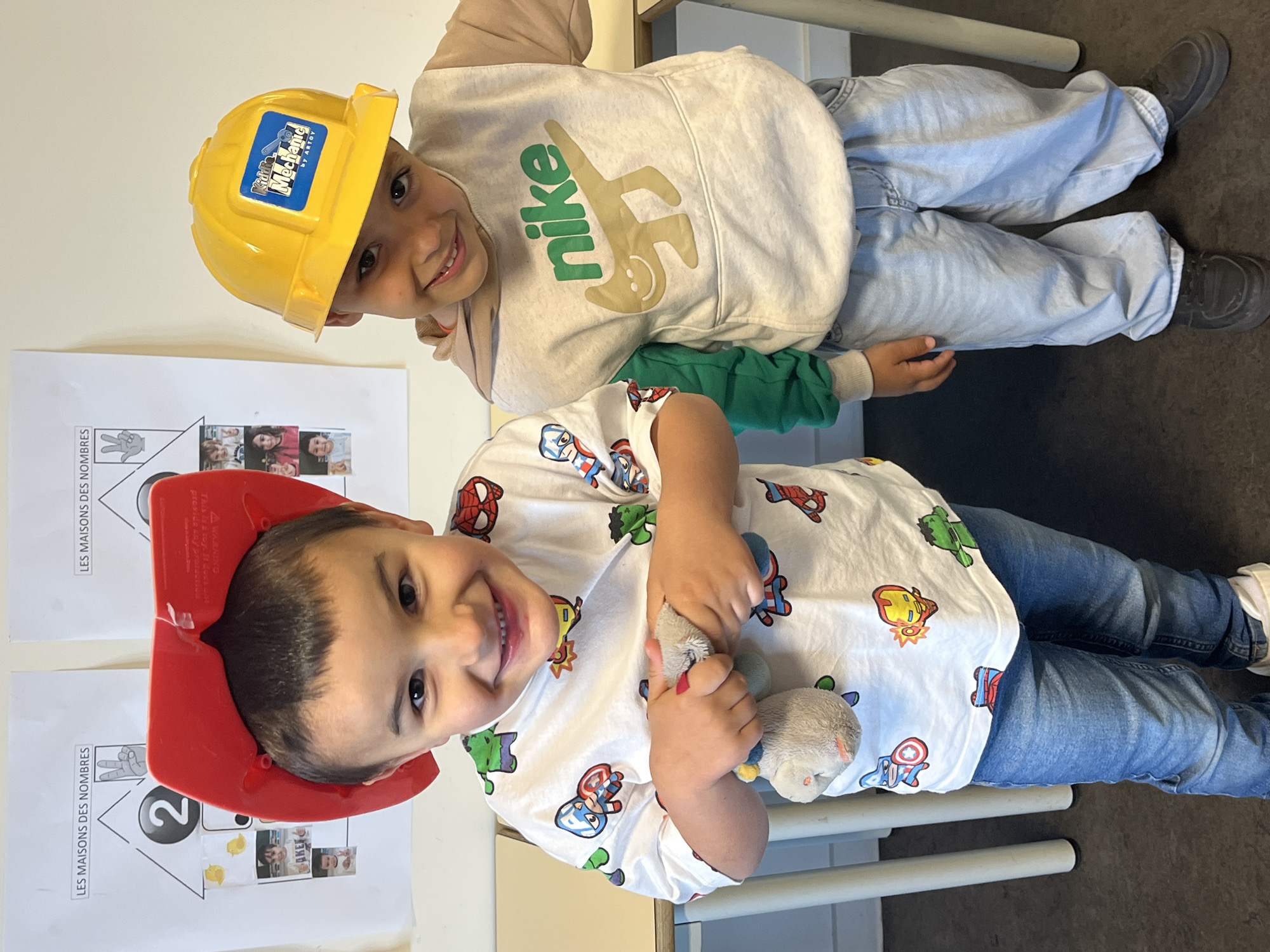 Two young boys are sitting on a table indoors. The boy in the foreground is wearing a white t-shirt with Marvel superhero imagery, blue jeans, and a red plastic construction helmet. He is holding a small stuffed animal. The boy in the background is wearing a beige sweatshirt with a green "Nike" logo, light blue jeans, and a yellow plastic construction helmet. Papers with French text and images are visible on the table next to them. The text includes "LES MAISONS DES NOMBRES" and "AWARNING". The photograph appears to have been taken in La Fouillouse, France.