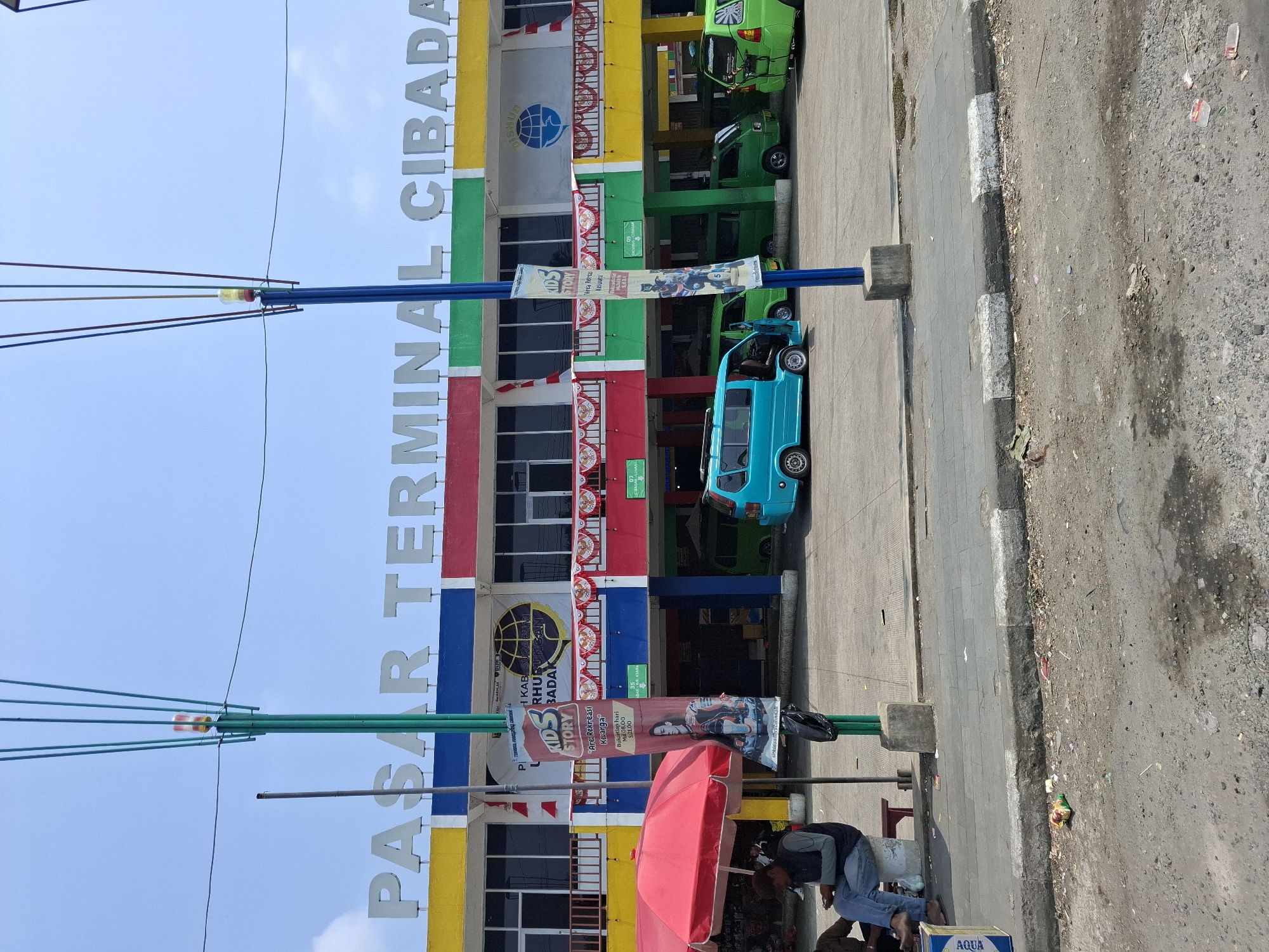 The image depicts the entrance to the Cibadak Terminal Market (Pasar Terminal Cibadak) in Cibadak, Indonesia. The building is brightly colored with sections of yellow, blue, red, and green. Large, white lettering above the entrance reads "PASAR TERMINAL CIBADAK". A tall, green pole with several wires extends upwards, topped by a blinking light.
In the foreground, a paved area with a curb separates the street from the sidewalk. Scattered debris, including plastic wrappers and small rocks, litter the ground. Several people are visible. One person, wearing a blue jacket and jeans, is sitting on a white circular object and appears to be looking down. Another person is partially visible to their left, also seated.
The terminal itself has multiple levels with windows and balconies. Red and white decorative banners are hung along the lower level. Several vehicles are parked in front of the building, including a bright turquoise minivan and several lime green vans, some of which appear to be public transportation. A red umbrella is set up to the left of the frame, providing shade. A black trash bag is tied to the green pole. A vertical banner advertising "KIDS STORY" with an image of a person in racing gear is hanging from the pole. The sky is a clear, pale blue.