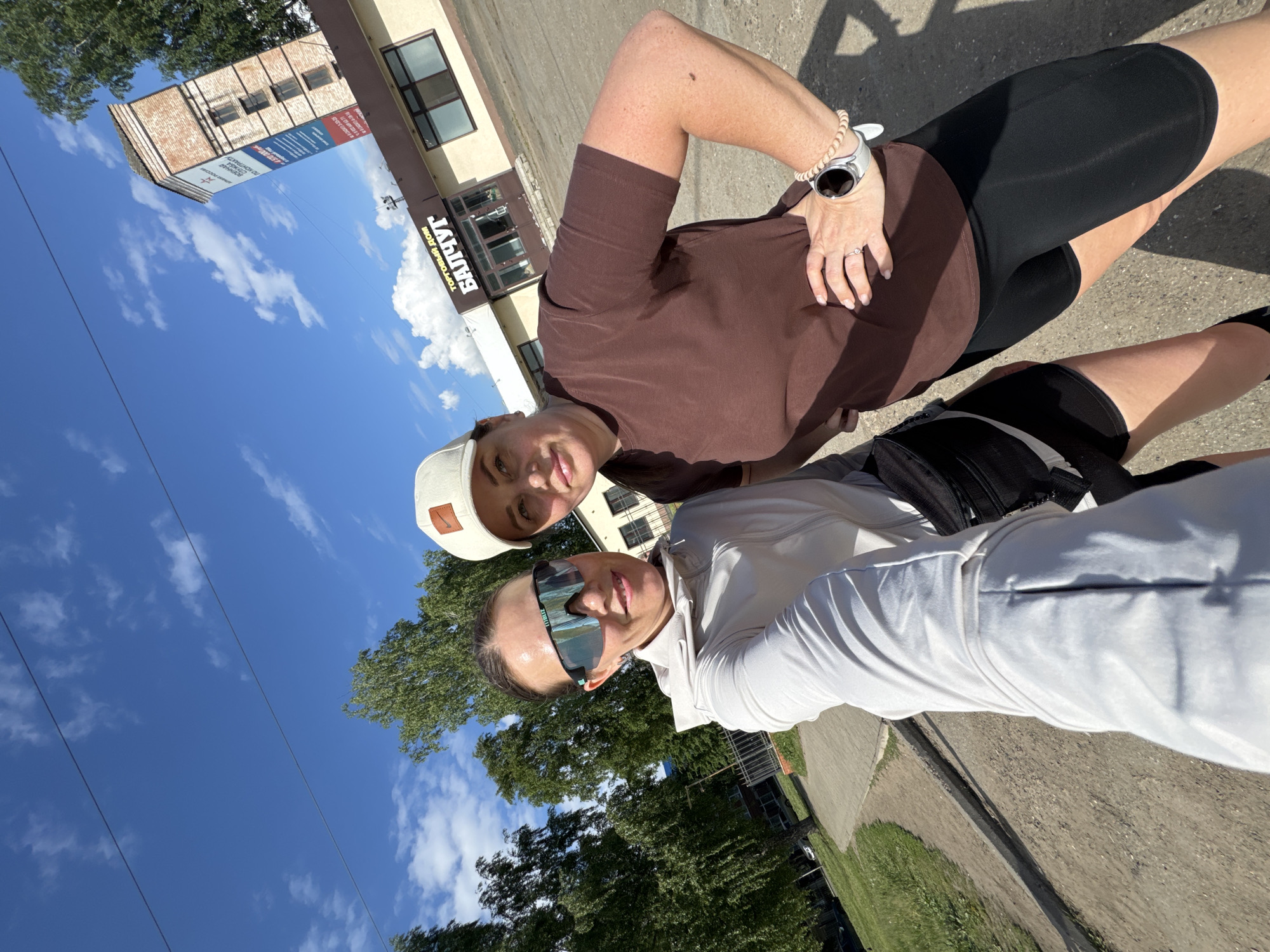 Two women are captured in a selfie outdoors on a bright, sunny day in Chaikovsky, Russia. The image is taken from a low angle, looking upwards towards the sky and buildings.
The woman closer to the camera, on the right, wears a light-colored (possibly white or light grey) long-sleeved top and large, sporty dark sunglasses. Her dark hair is pulled back. The second woman, slightly behind and to the left, wears a short-sleeved brown top, black form-fitting shorts (possibly cycling shorts), and a light beige baseball cap with a dark brown Nike swoosh logo patch on the front. She has a white smartwatch on her left wrist and an engagement ring on her left ring finger. Both women are looking towards the camera, with a slight smile on their faces, suggesting a casual and relaxed mood.
The background reveals a clear blue sky with scattered white clouds and lush green trees. Several buildings are visible. One building, directly behind the women, has a sign with Cyrillic text, prominently displaying "ТОРГОВЫЙ ДОМ" (Trading House) and "БАЙКУТ." Below this, on a blue section, additional Cyrillic text is present, including "Рекламное агентство" (Advertising agency). Further in the background, another brick-faced building with multiple windows can be seen, along with a large billboard featuring more Cyrillic text, with "РОДНАЯ" (Native/Homeland) partially visible. Power lines stretch across the sky. The ground appears to be a paved surface, likely a sidewalk or road, with some grassy areas nearby. The scene suggests an urban or suburban setting.