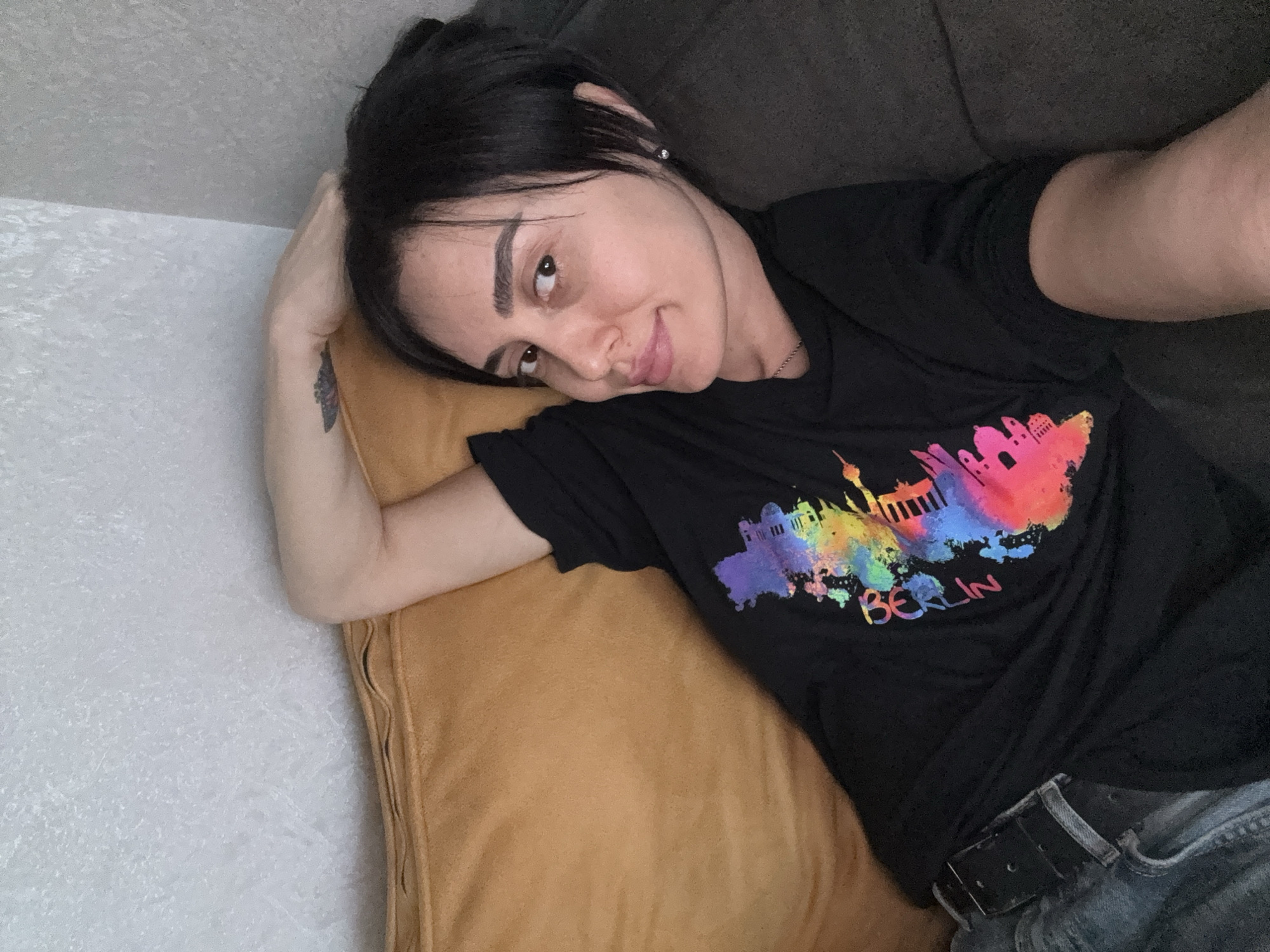 A woman with dark hair and fair skin is lying on a mustard-colored pillow, positioned against a gray couch. She is wearing a black t-shirt with a colorful graphic of the Berlin skyline and the word "Berlin" printed beneath it. She is also wearing a pair of jeans and a black belt. Her head is tilted towards the camera, and she has a slight smile. Her left arm is bent and resting near her head on the pillow, and there is a small tattoo on her left forearm. The background consists of a textured, light gray wall. The scene does not depict any elements indicating a location in Tbilisi, Georgia.