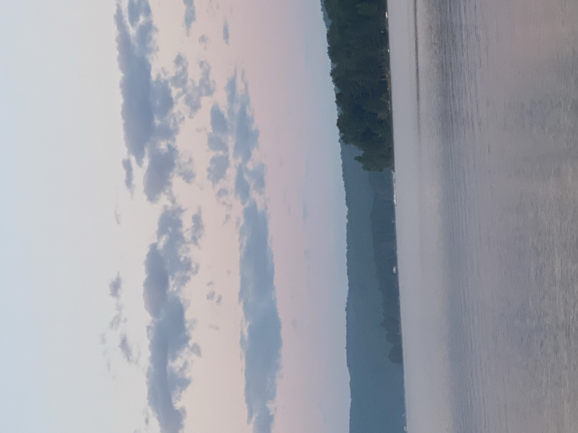The image depicts a serene lake scene, likely at dusk or dawn, in the vicinity of Duhamel, Canada. The water is calm with a slight mist rising from its surface, creating a soft, ethereal effect. In the distance, a tree-covered shoreline forms a dark silhouette against a sky transitioning from pale blue to soft pinks. Scattered, wispy clouds are visible in the upper portion of the frame. The overall atmosphere is tranquil and peaceful, characteristic of a natural lakeside environment. No people or specific structures are clearly discernible, emphasizing the natural landscape.