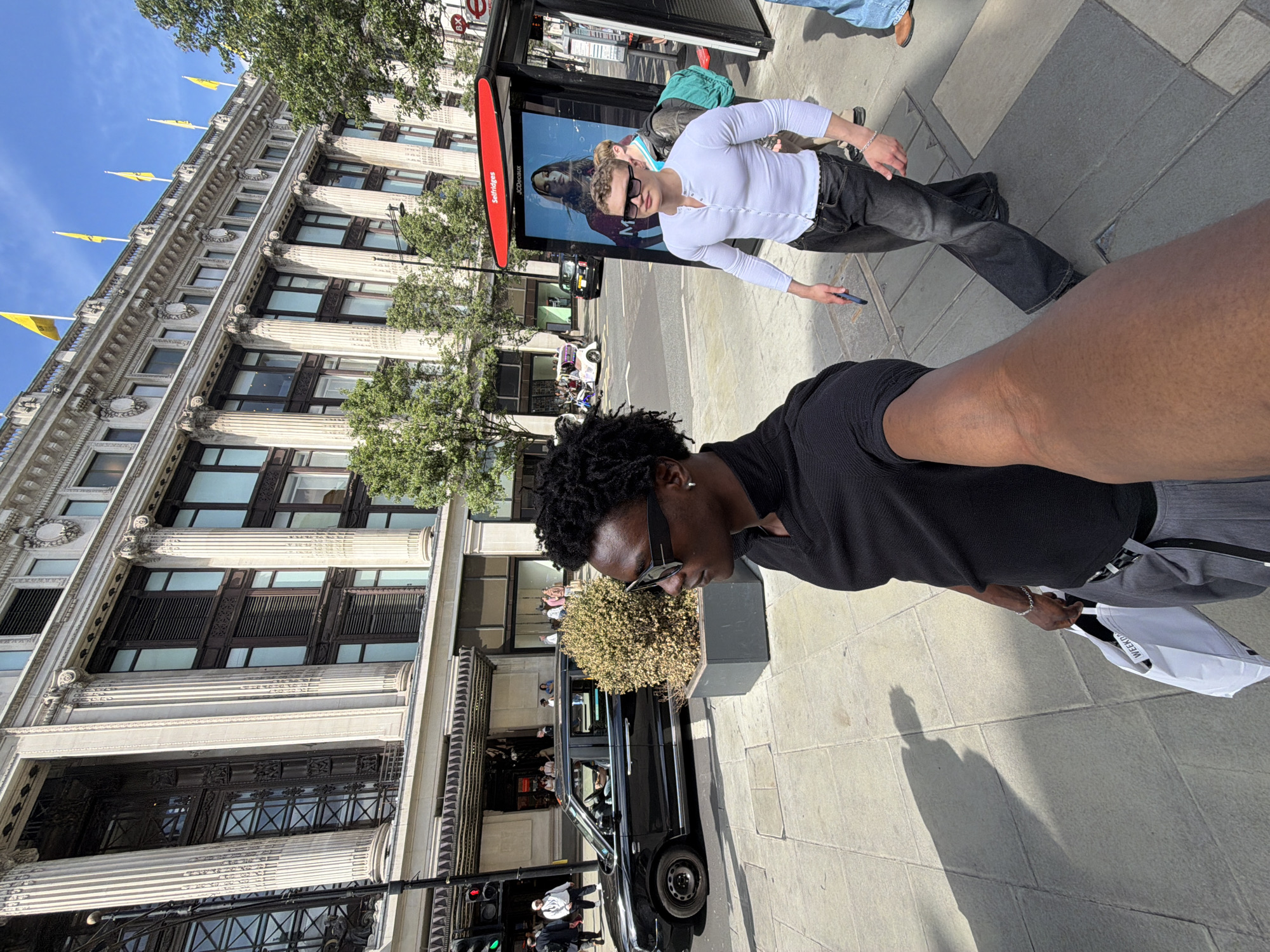 Here is a description of the image:
The image shows two men taking a selfie on a street in London, United Kingdom. The man taking the photo is wearing a black shirt and sunglasses and is positioned on the right of the image. Next to him, another man wearing a white shirt and dark sunglasses is looking at the camera while holding a phone. They are standing in front of a bus stop and a black taxicab. In the background is the Selfridges building, adorned with yellow flags. The sidewalk is visible, as well as other pedestrians walking in the area. The lighting suggests it is daytime.