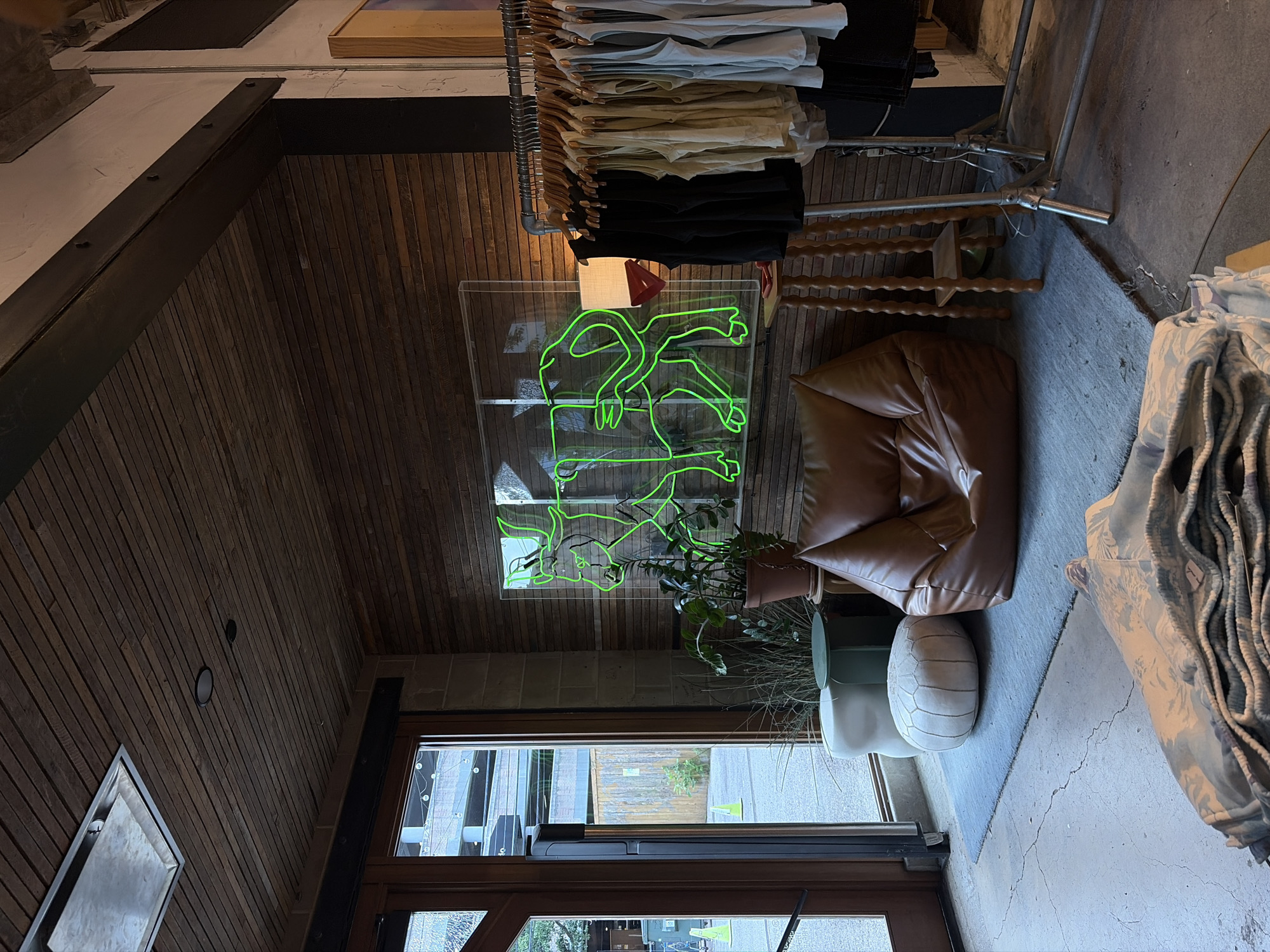 The photo shows the interior of a retail space, likely a clothing store, with a rustic aesthetic. The walls are clad in horizontal wooden planks and the ceiling features exposed beams and ductwork. A clothing rack loaded with garments in neutral tones and black occupies a portion of the upper right. Below the rack, a transparent sign featuring a neon-green graphic of an animal in a playful pose is visible. A brown leather beanbag chair is positioned near the sign. A potted plant sits on the floor, adjacent to a smaller, cream-colored pouf. A full-length glass door allows a view of an exterior area with green foliage and urban elements. The location may be Austin, Texas, based on the image's context within a dataset.