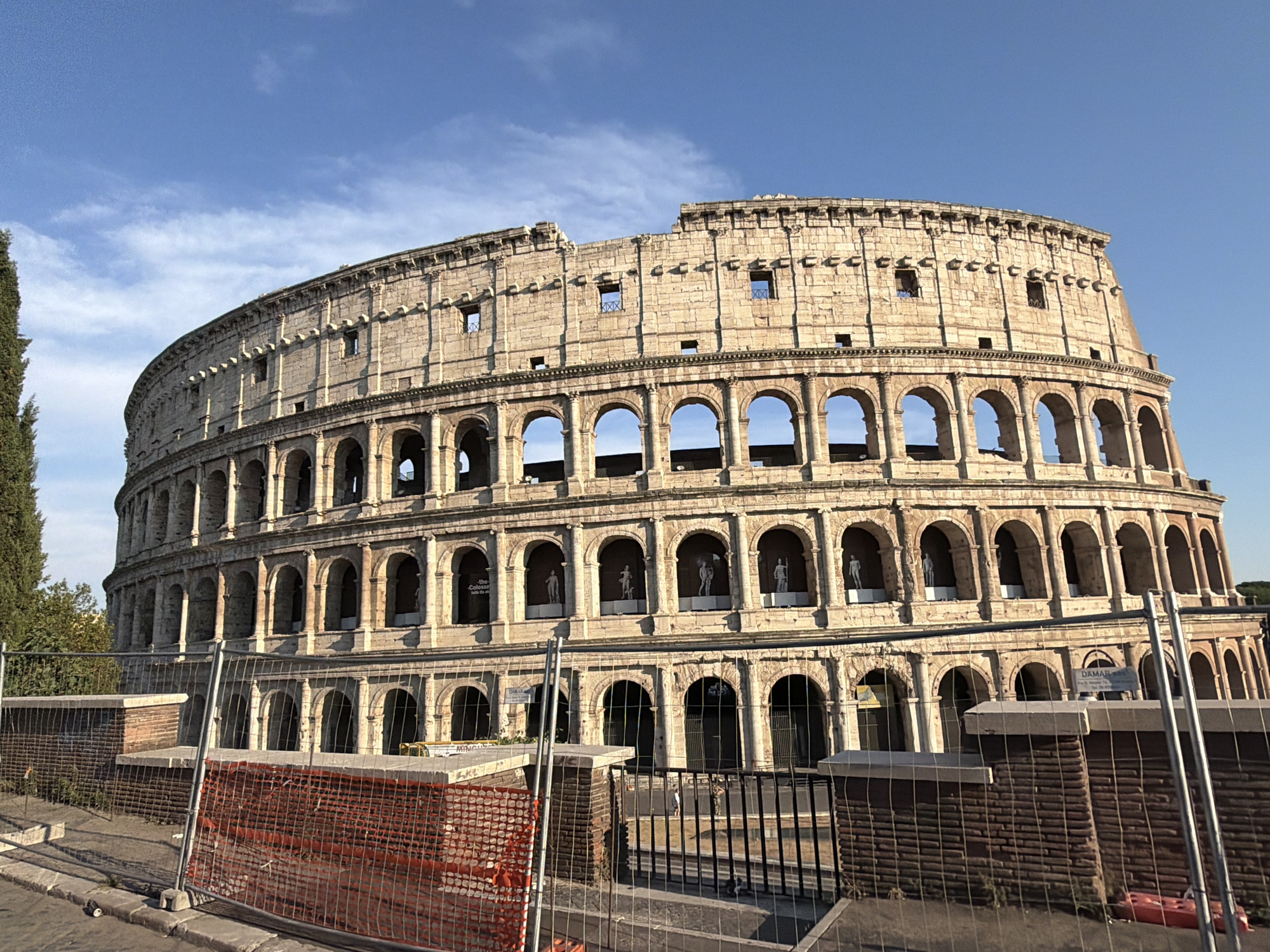 The image shows a sunlit view of the Colosseum in Rome. The ancient amphitheater is made of weathered stone and features multiple tiers of arches. In some of the upper arches, there are white statue-like figures visible. The sky above is a clear, bright blue with a few wispy clouds.
In the foreground, there are temporary construction fences with orange netting and black metal bars, suggesting ongoing work or preservation efforts. Some brickwork is visible behind the netting. The lighting indicates it is daytime, likely late morning or early afternoon, as the sun casts distinct shadows on the structure. A tall, slender green tree stands to the left of the Colosseum.
There are no people clearly visible in the image, though the statues within the arches might be mistaken for people at first glance. The scene is one of historical grandeur contrasted with modern temporary barriers.
Visible text includes signs on the fence, one of which appears to say "DAMAR" and another, further to the right, has some text that is too blurry to read clearly, but it looks like "THE COLOSSEUM".