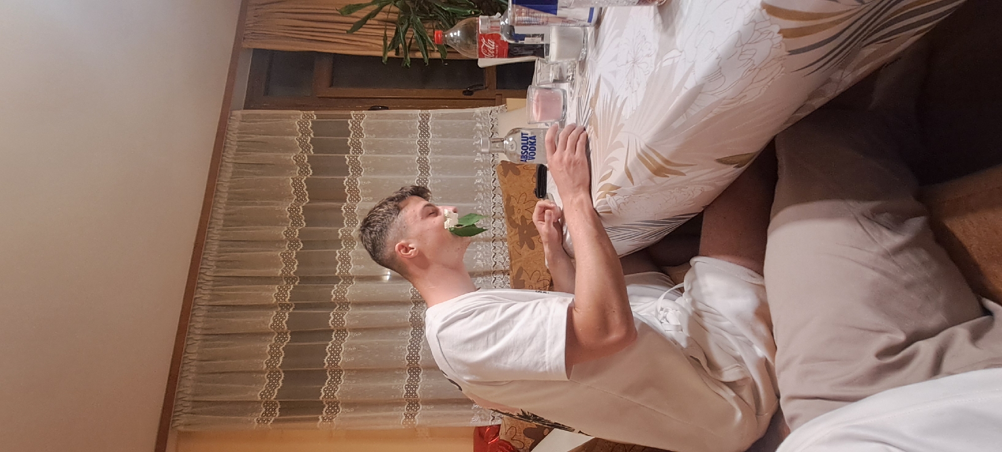 A young man is sitting at a table in a room. He has a white flower and green leaves in his mouth. The table is covered with a white tablecloth with a floral pattern. There are several glasses and bottles of alcohol on the table, including Absolut Vodka and Coca-Cola. In the background, there is a window with lace curtains. The location could be Burgas, Bulgaria, but there is no concrete information to prove the location.