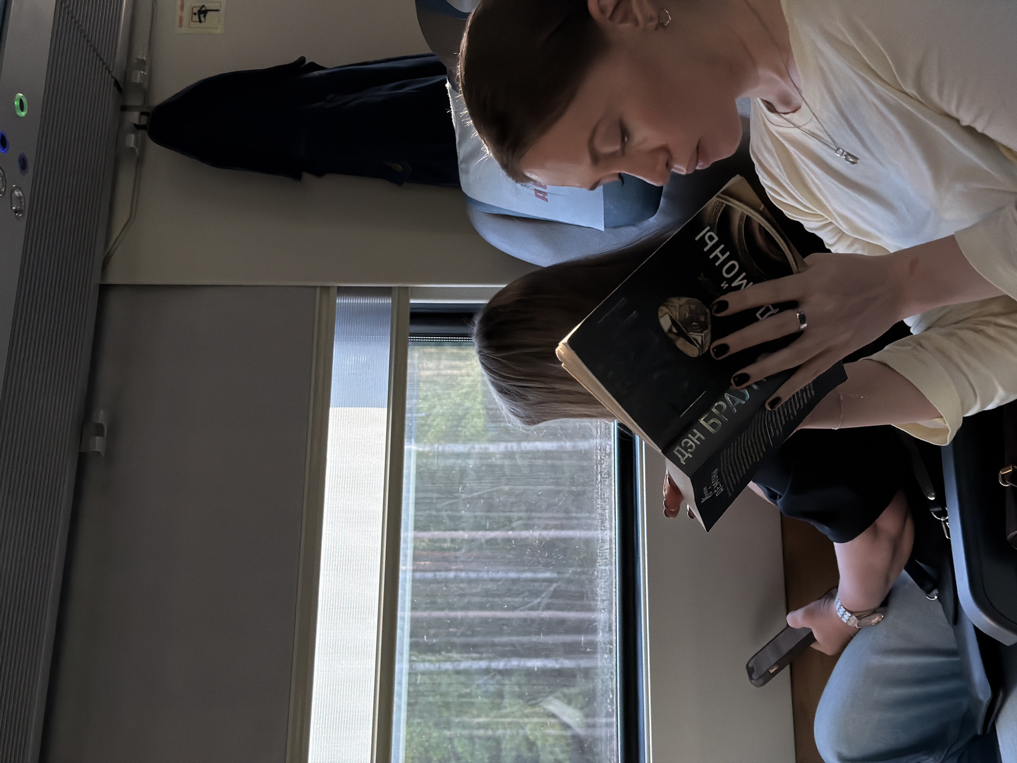 Inside a train carriage, two women are seated by a window. The first woman, with her hair pulled back, is holding a book with a dark cover and text in a Cyrillic alphabet, with the name "Дэн Браун" visible. She is wearing a light-colored top, a necklace, and rings on her fingers, and her nails are painted black. The second woman is also reading, and holding a smartphone in her hand. The train window shows a blurry view of trees passing by. On the left, there's a section of the train interior with control buttons. The overhead space contains a hanging item, possibly a jacket or bag. The location is Ушаки, Russia, based on the visible text on the book.