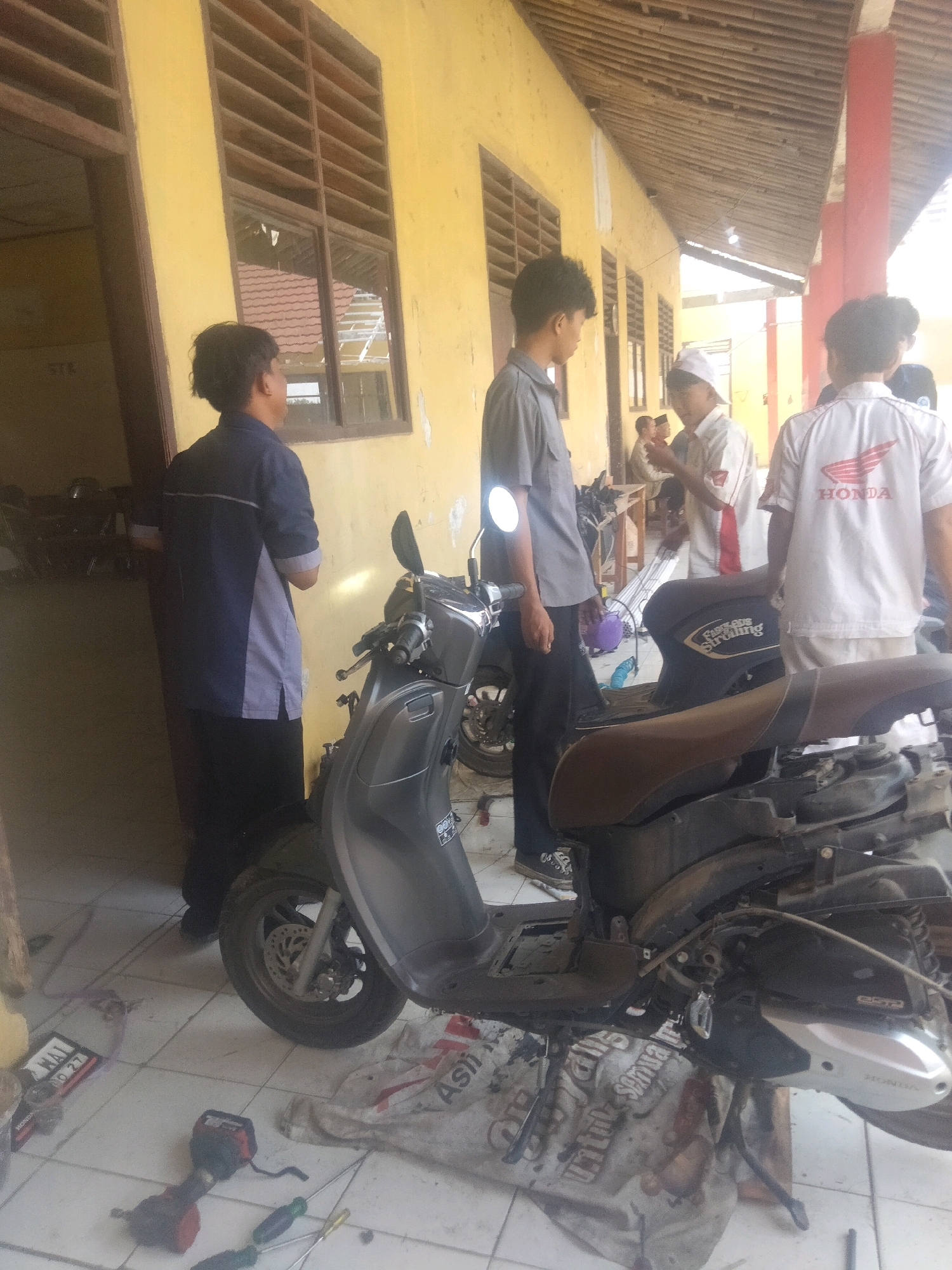 A group of young men are gathered outdoors in what appears to be a schoolyard or workshop area. The building in the background has yellow walls and wooden window louvers. A dark gray scooter is parked on the tiled ground, and it appears to be undergoing some form of maintenance or repair, with parts exposed. One man on the left, wearing a blue and gray shirt and black pants, stands near the scooter. To his right, another young man in a gray shirt and black pants is also near the scooter. Further back, a man in a white shirt with red accents and a white cap is visible, along with another person in a white shirt with "HONDA" logo visible on the back. Tools, including a red drill and screwdrivers, are scattered on the ground around the scooter. The overall scene suggests a casual, practical setting, possibly a vocational training area or a communal space for maintaining vehicles. The location is identified as Suka Sari, Indonesia.