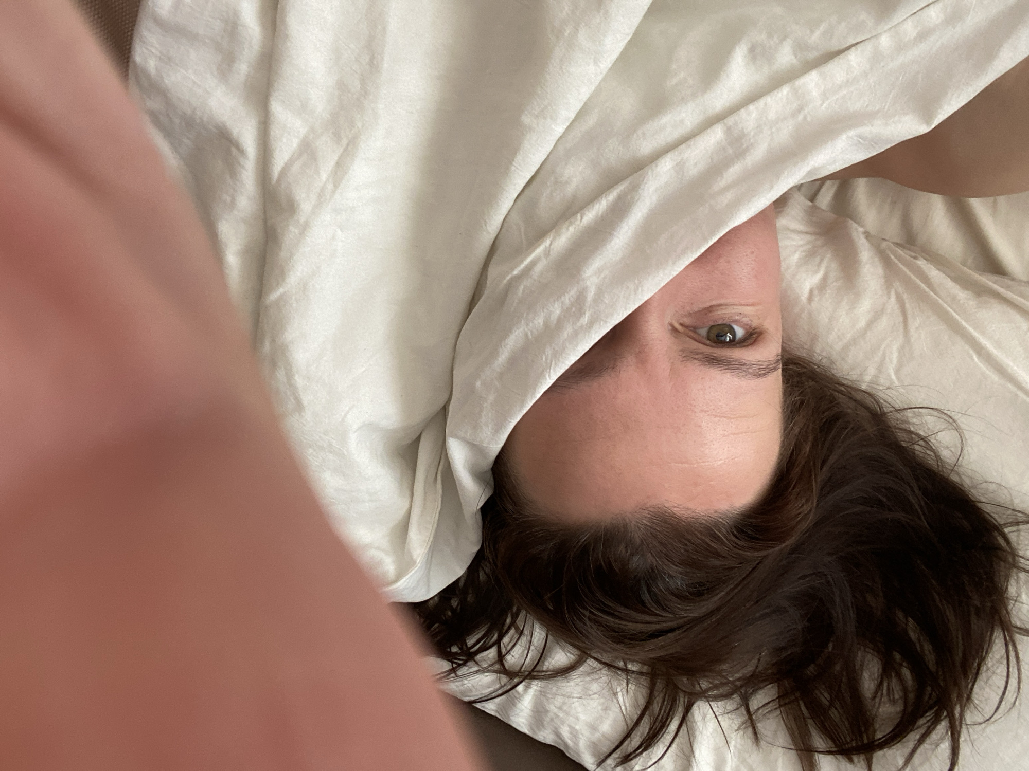 A person is lying in bed, partially covered by a white sheet. Only one eye is visible, looking directly at the camera. The person has dark brown hair. A blurred object, possibly a hand or arm, is in the lower left of the frame. The setting appears to be a bedroom. The location is unknown, and there is no evidence of Chaikovsky, Russia.