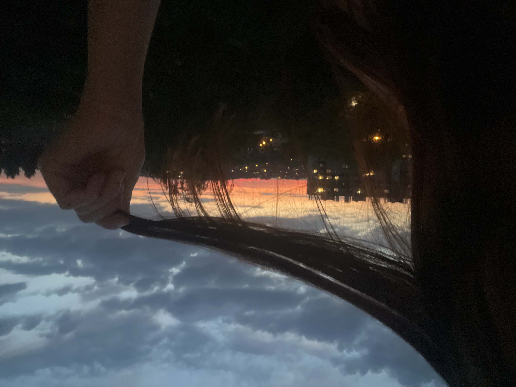 A close-up, low-angle shot focuses on a person's dark hair and hand against a backdrop of a city skyline at dusk. The hair, obscuring the left side of the frame, is held by a right hand which is partially visible in the lower right corner, gripping a lock of hair. In the mid-ground, a hazy city panorama unfolds with buildings illuminated by warm lights, and trees silhouetted against the vibrant orange and pink hues of the sunset sky. Dark, scattered clouds populate the upper portion of the image. The overall lighting is dim, characteristic of twilight, with the sky providing the most significant illumination. The location is noted as Unknown, Unknown.