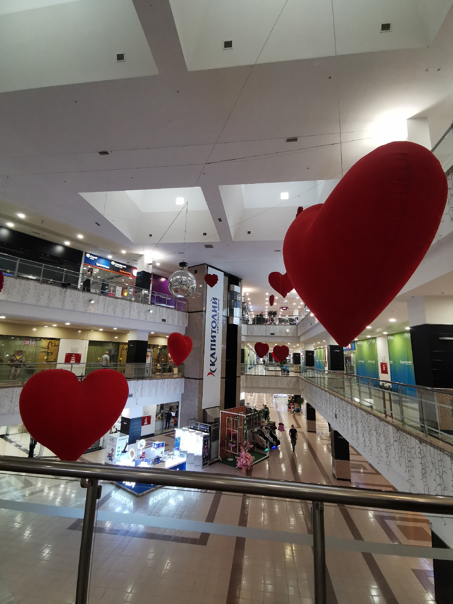 The image shows the interior of a multi-level shopping mall, possibly in Moscow, Russia, based on the "КАПИТОЛИЙ ТОРГОВЫЙ ЦЕНТР" sign visible. The mall is decorated with large, red heart-shaped balloons suspended from the ceiling, suggesting a Valentine's Day theme. The floors are tiled, and there are several store displays visible on different levels. People are seen walking throughout the mall. The architecture includes multiple levels with railings and open spaces. A disco ball hangs from the ceiling.