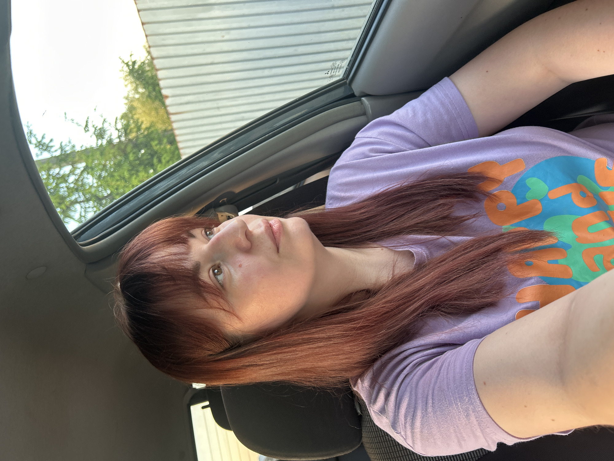 A woman with auburn hair and a lilac t-shirt is taking a selfie inside a car. The car has an open sunroof, through which trees and a building can be seen. The location cannot be determined to be Фоки, Russia.