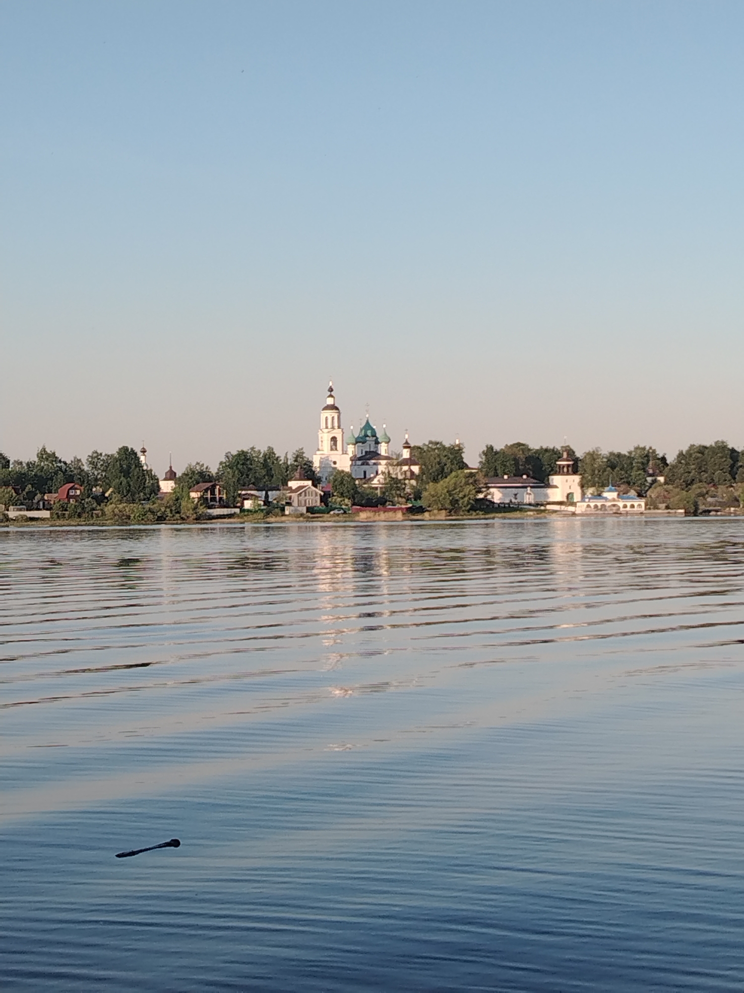 The image shows a water body with buildings visible in the distance. The buildings appear to be part of a settlement or town, with prominent structures that suggest churches or religious sites. The water surface has gentle ripples, indicating a slight breeze or movement on the water. A stick or small object is floating in the water in the foreground. The sky is clear and light blue. This is likely Rusanovo, Russia.
