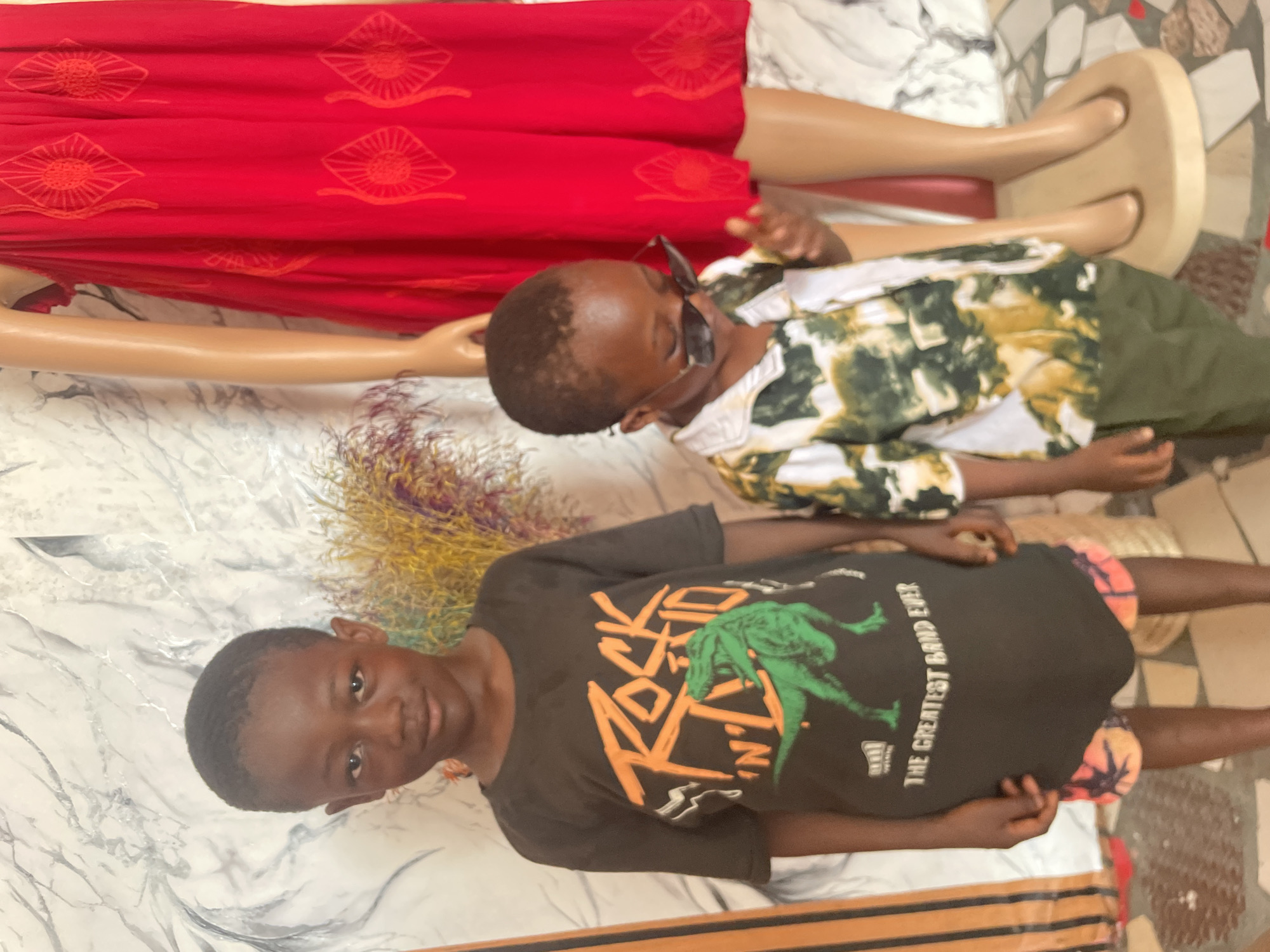 The image depicts two young boys in what appears to be a retail or display setting. One boy, wearing a black t-shirt with a "ROCK" graphic and a green dinosaur, is positioned in the foreground and looks directly at the camera. The second boy, wearing sunglasses and a patterned shirt with green pants, is in the background partially obscured by a mannequin with a red dress.
The setting includes what seems to be a mannequin dressed in a red dress. The presence of the mannequin suggests this location is potentially a clothing shop or display area. The flooring appears to be marble with brown accents.
Without further information, a specific location within Badagry, Nigeria, cannot be precisely determined.
