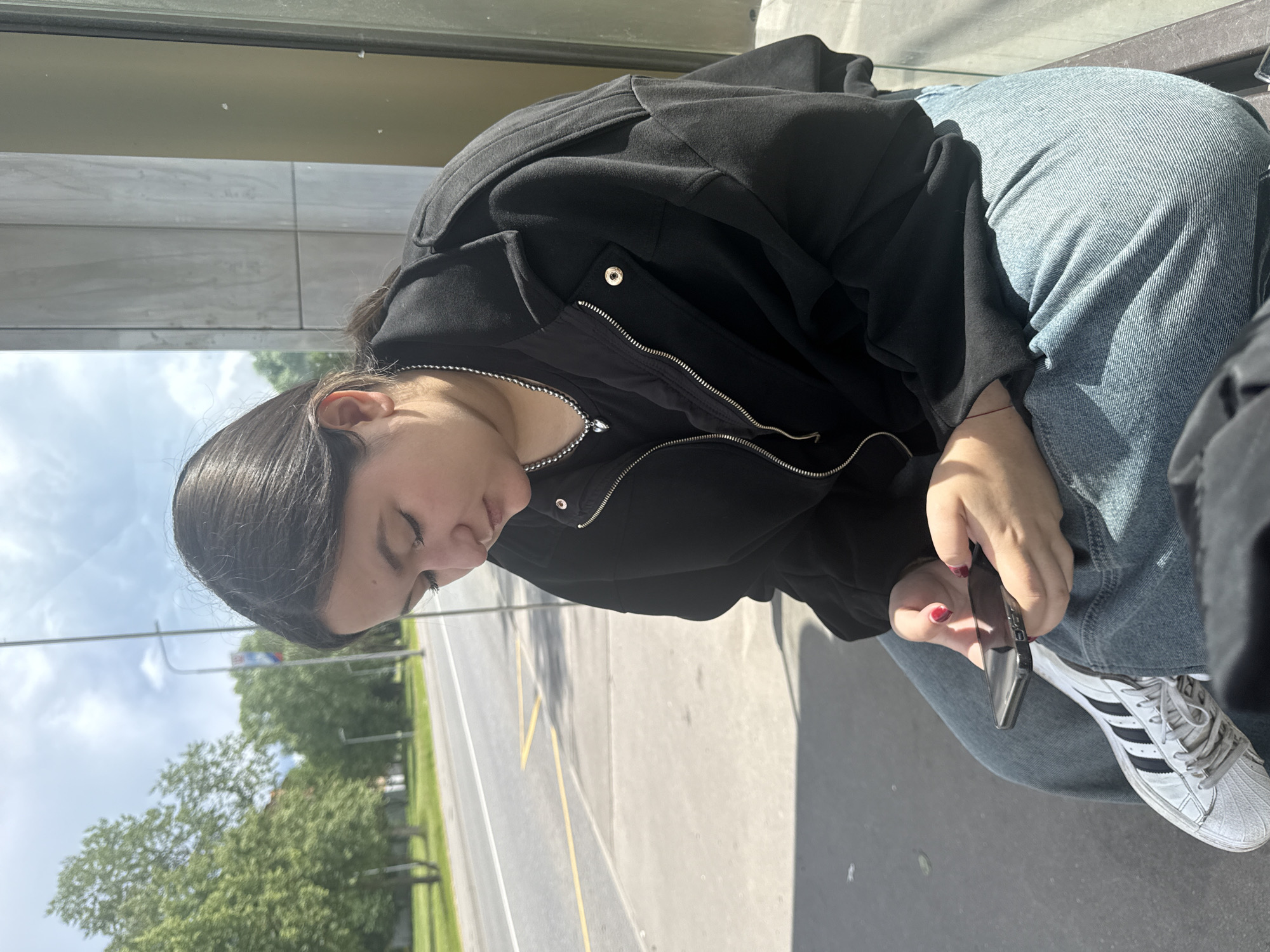 A young woman with dark hair is sitting outside, looking down at her phone. She is wearing a black jacket and blue jeans. Her fingernails are painted red. She is wearing white sneakers with black stripes. In the background, there is a street with cars, trees, and a bus stop sign. The weather appears to be sunny. The location is Yakovlevskoye, Russia.