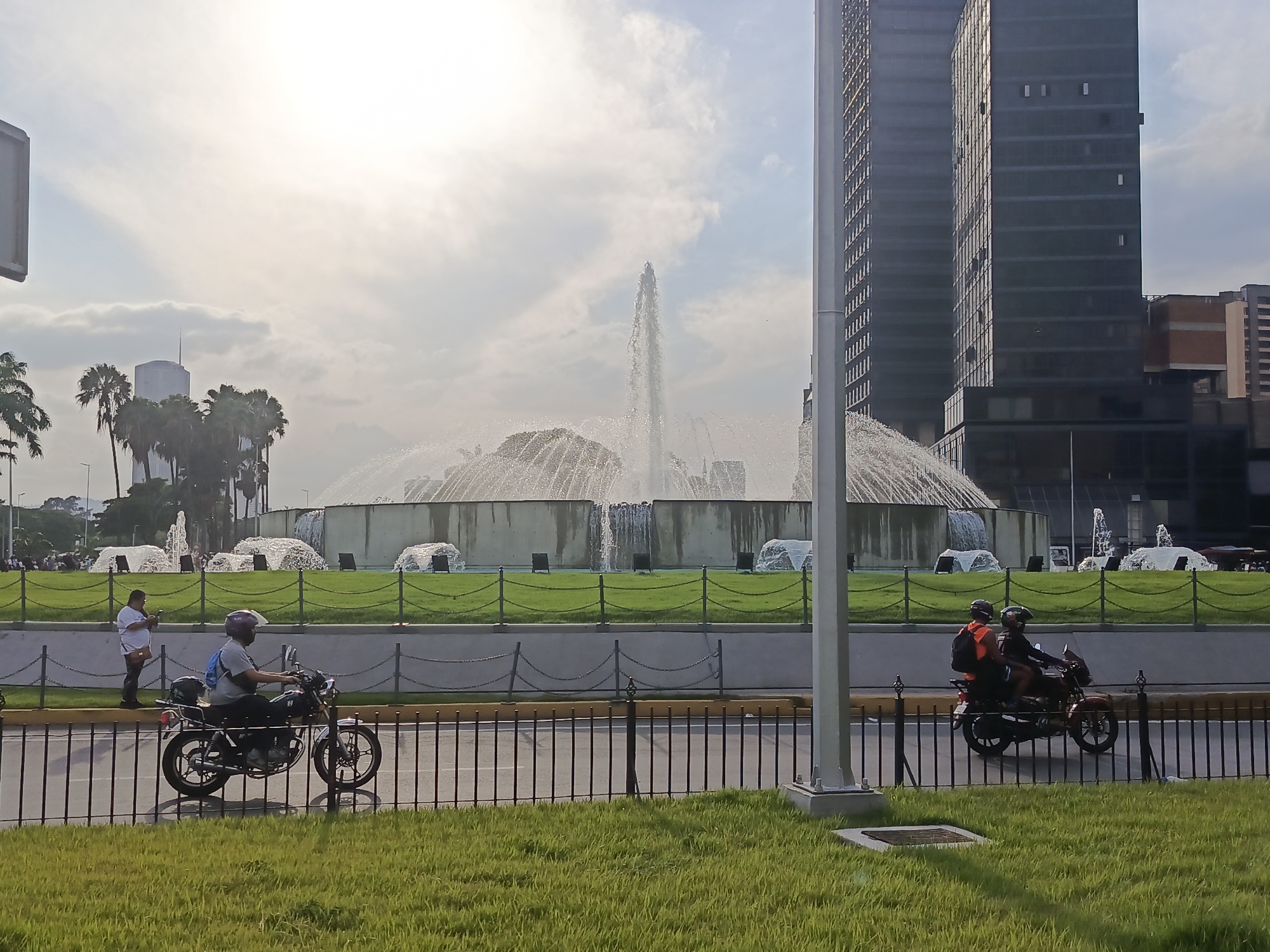 This media file presents a bright, sunny urban landscape in Caracas, Venezuela, centered around a large, active fountain. The time of day appears to be midday or early afternoon, with intense sunlight creating some glare in the upper left of the frame and illuminating the scene.
The dominant subject is a grand, circular fountain with multiple water jets, including a tall central spray and numerous smaller cascades around its concrete base. This fountain is surrounded by well-maintained green grass, which also forms median strips in the foreground.
In the background, modern architecture defines the cityscape. To the right, several tall, dark skyscrapers with reflective windows stand prominently. Further to the left, behind a cluster of green palm trees, a distinctive light-colored cylindrical building with a spire rises into the sky. The sky above is bright with scattered clouds.
The foreground is separated from the fountain area by a black metal fence with chain links. On the paved street to the left, a person wearing a helmet and a blue backpack is seated on a black motorcycle, seemingly stopped, while another individual stands next to it. On the right side of the street, two people, both wearing helmets, are riding a red motorcycle, indicating movement through the urban environment. The driver of the red motorcycle is wearing an orange safety vest. A large, light-colored pole, likely a utility or lamppost, bisects the right side of the image. The overall scene captures a vibrant moment of daily life in the city. No visible text is discernible in the image.